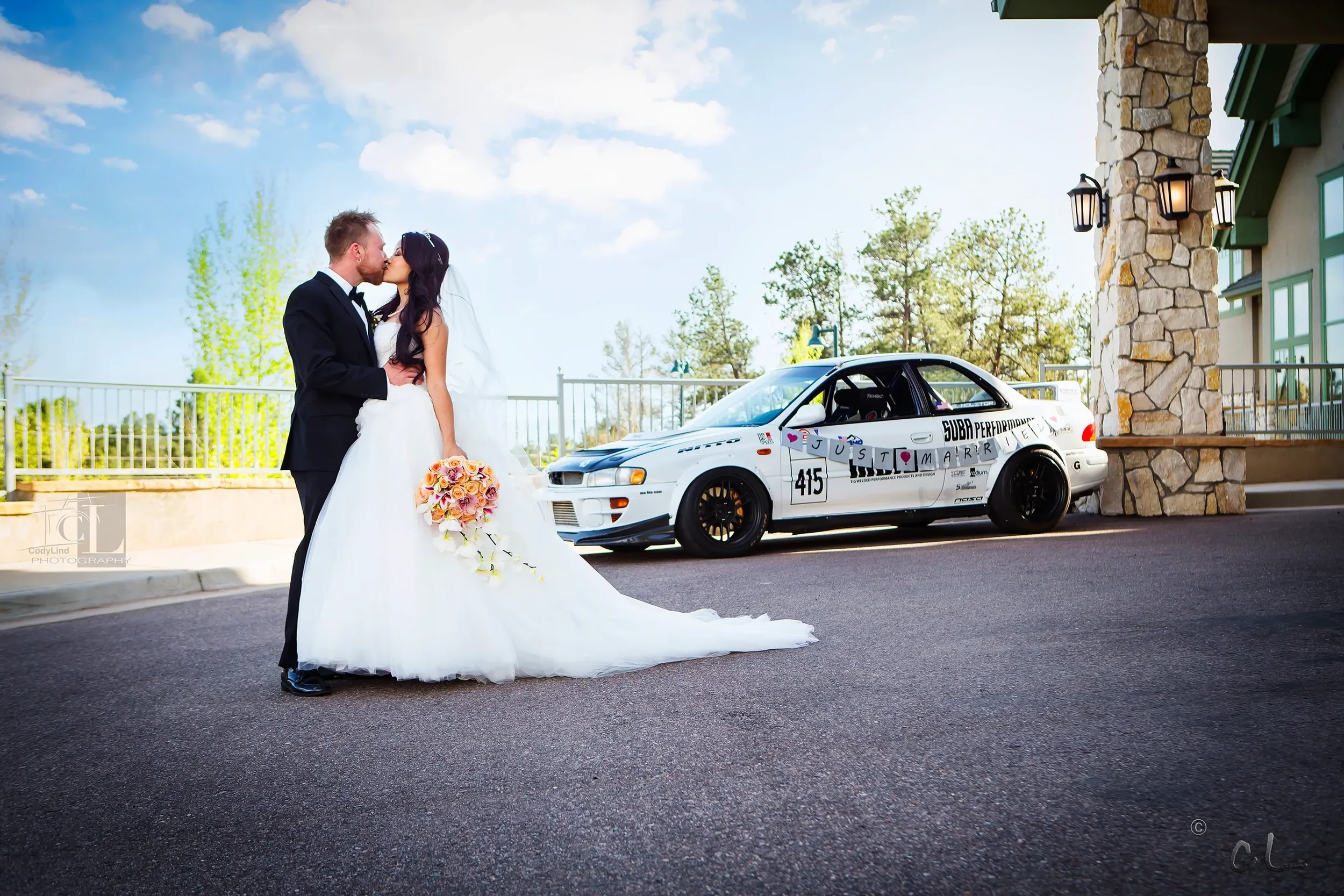 Bride and groom in wedding attire kissing in front of a rally car with racing decals, parked outside a building with stone and green accents, during daytime with a partly cloudy sky.