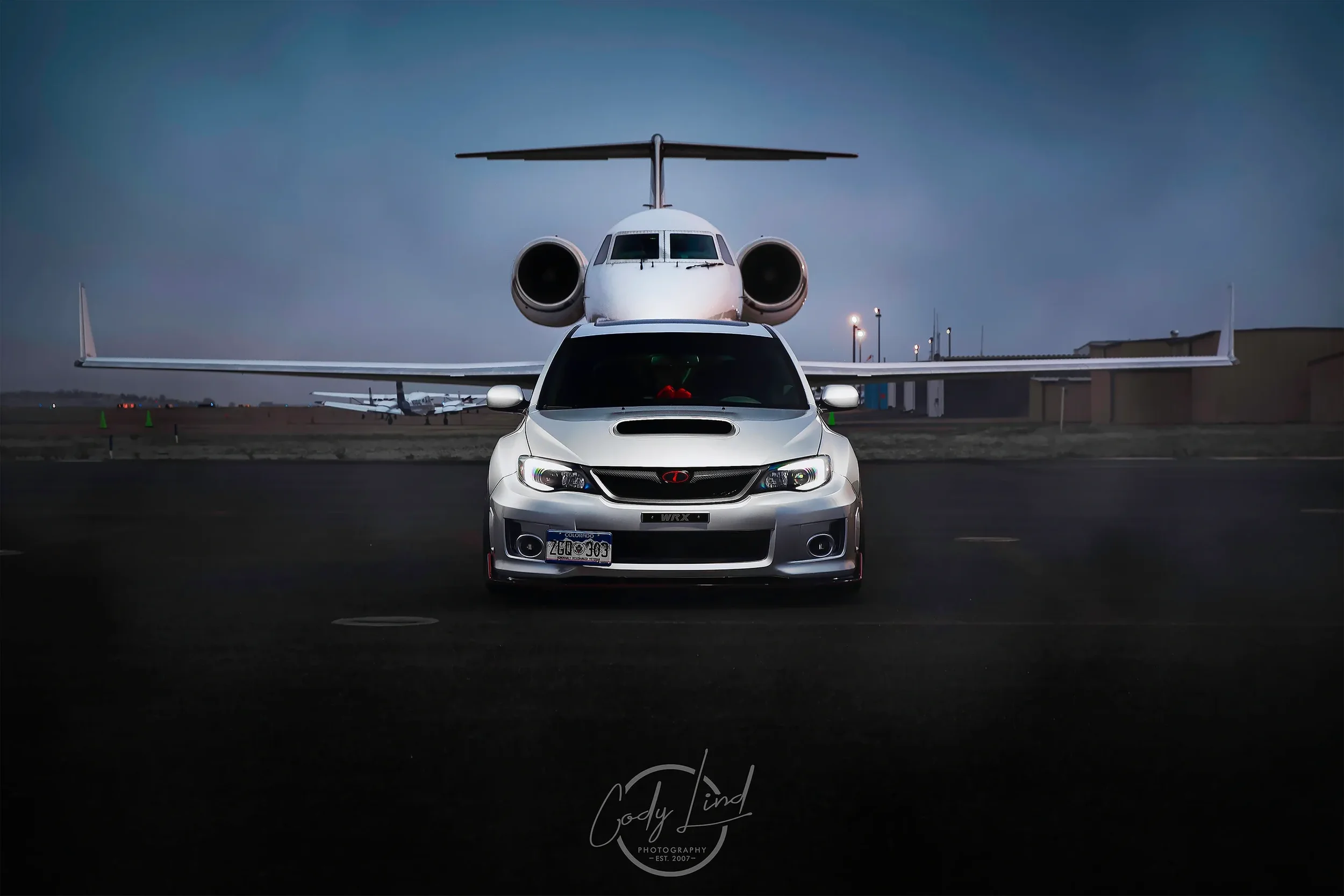 A silver car with a sporty design and a Subaru logo on the front, parked in front of a private jet on an airport tarmac during dusk.