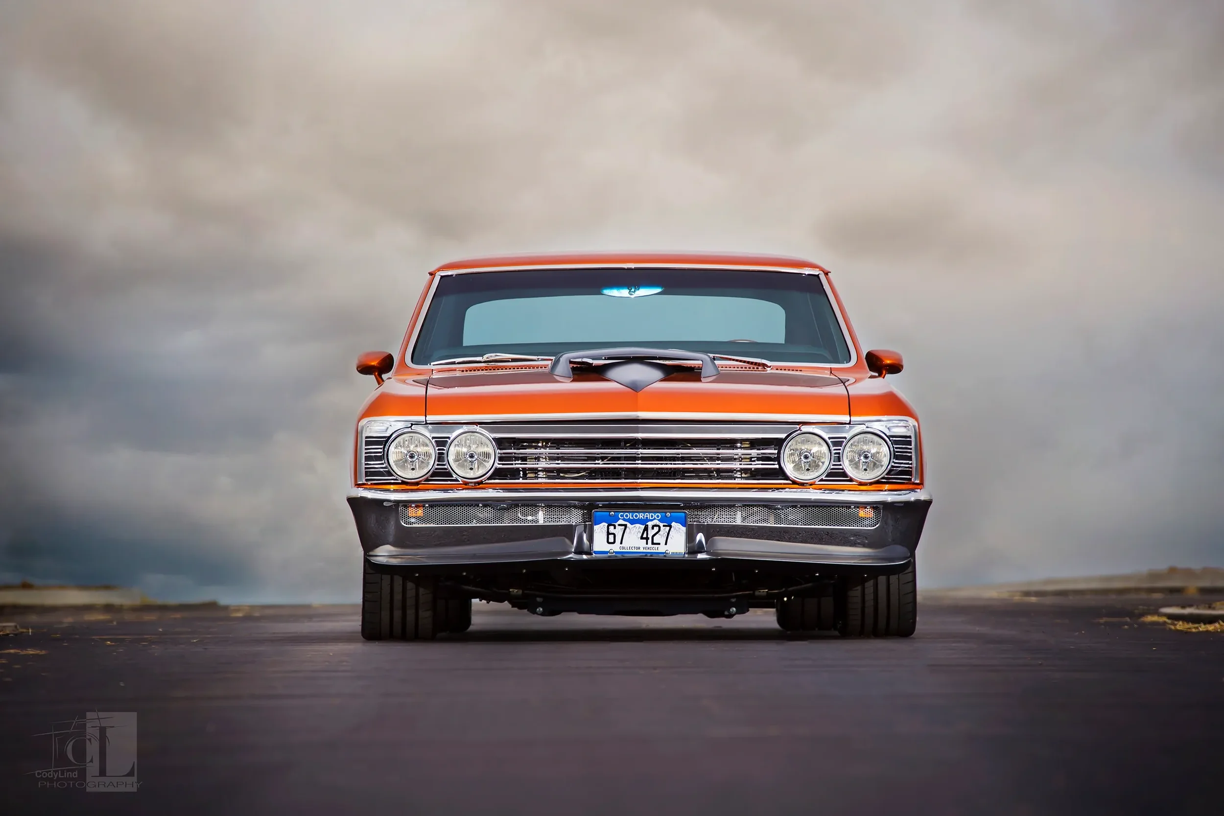 Front view of a vintage orange muscle car with a custom hood scoop, chrome grille, round headlights, and a Colorado license plate, parked on a paved road under a cloudy sky. 67 chevelle 