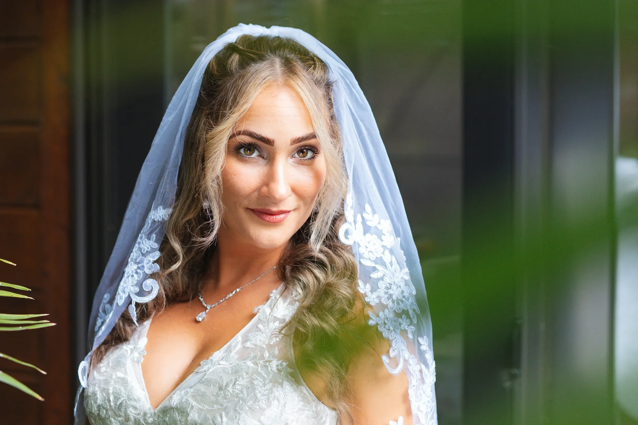 A bride with blonde hair and light makeup, wearing a veil and a white lace wedding dress, standing indoors with a dark background and some greenery in the foreground.