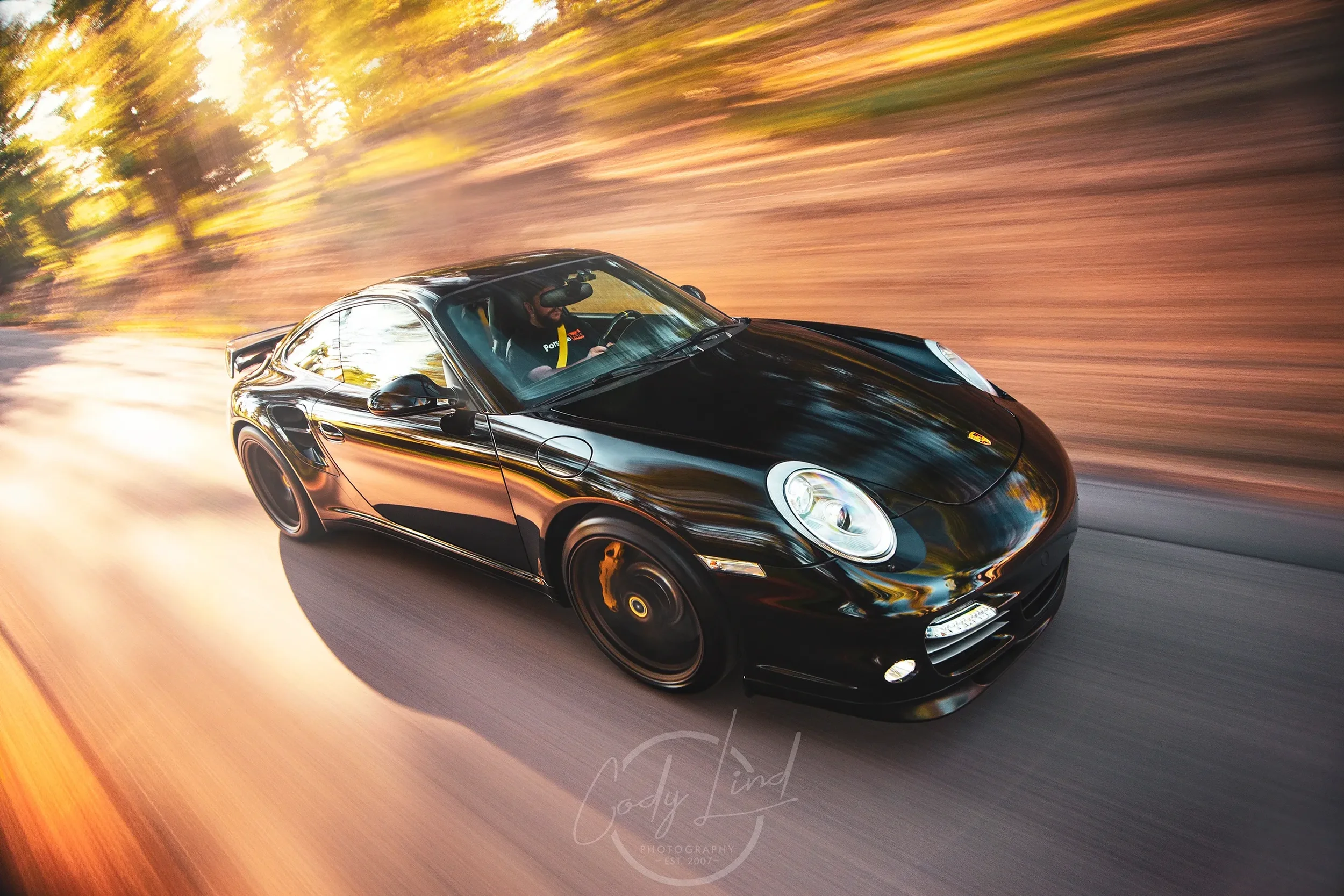 A black sports car, likely a Porsche, driving on a road surrounded by trees with sunlight filtering through the leaves, creating a blurred background indicating high speed.