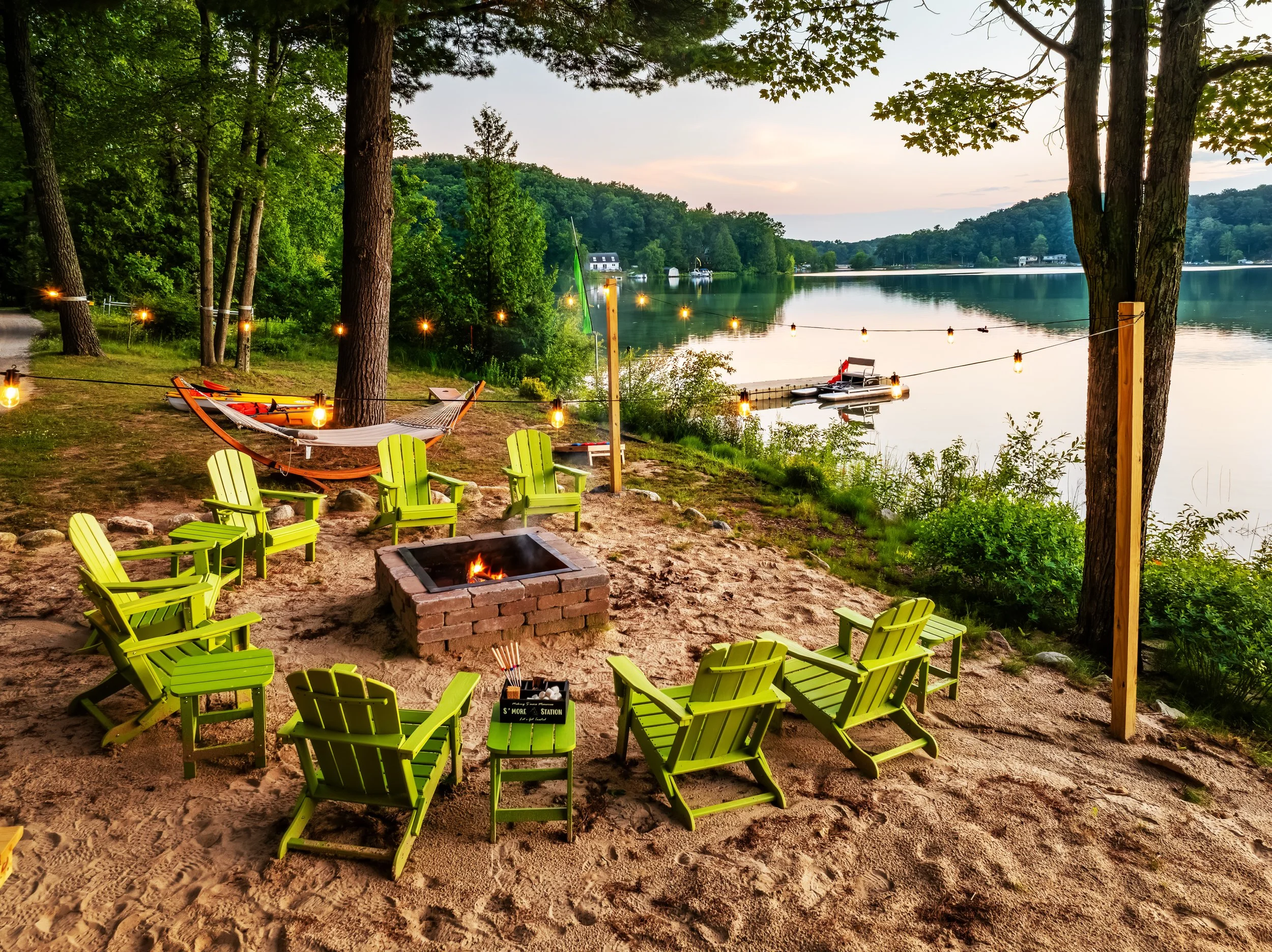 A lakeside outdoor gathering area with lime green Adirondack chairs arranged around a fire pit, string lights hanging between trees, a hammock behind the chairs, and a dock with boats on a calm lake.