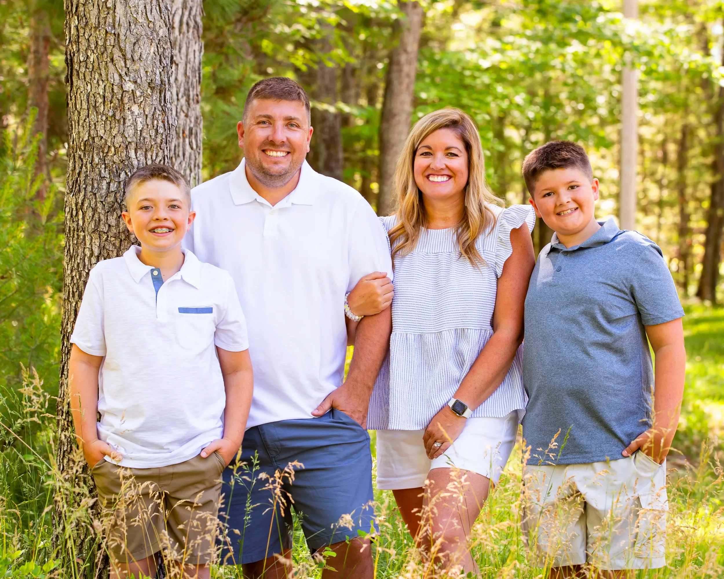 A smiling family of four outdoors in a wooded area. A woman holds hands with a man, with two boys standing beside them, all dressed in casual summer clothing.