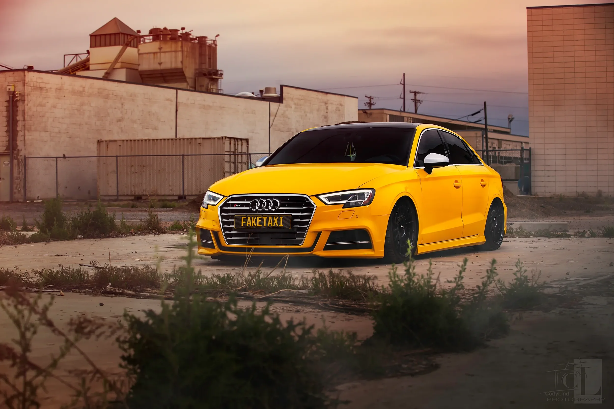 A yellow Audi car with a fake taxi license plate parked on an urban dirt lot.