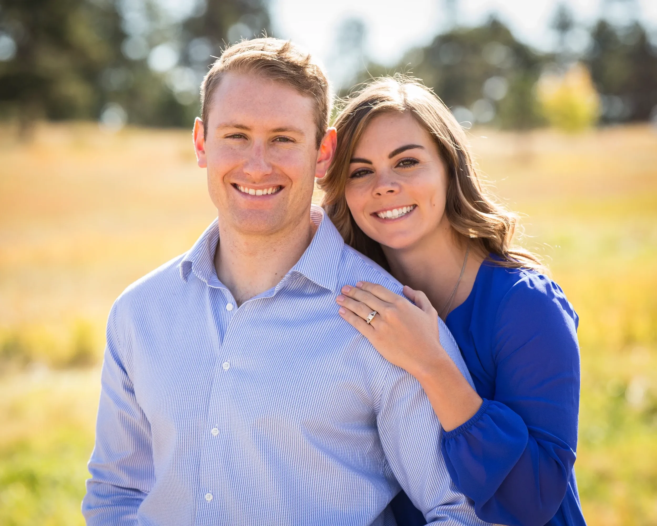 A smiling couple standing outdoors in a field with yellowish grass and trees in the background. The woman has her hand on the man's shoulder and is wearing a ring.