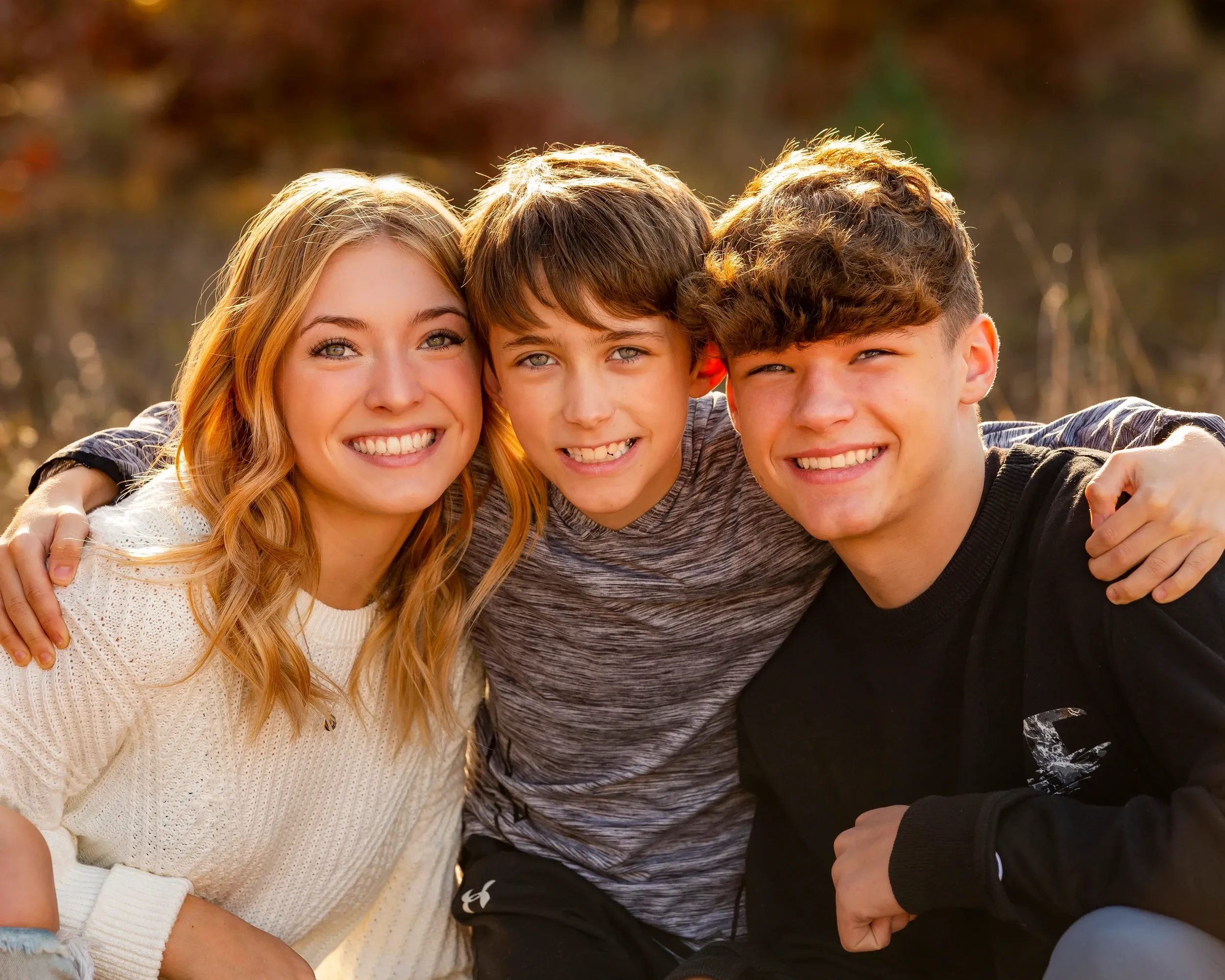 Three teenagers, two boys and one girl, smiling and hugging outdoors in a park during autumn.