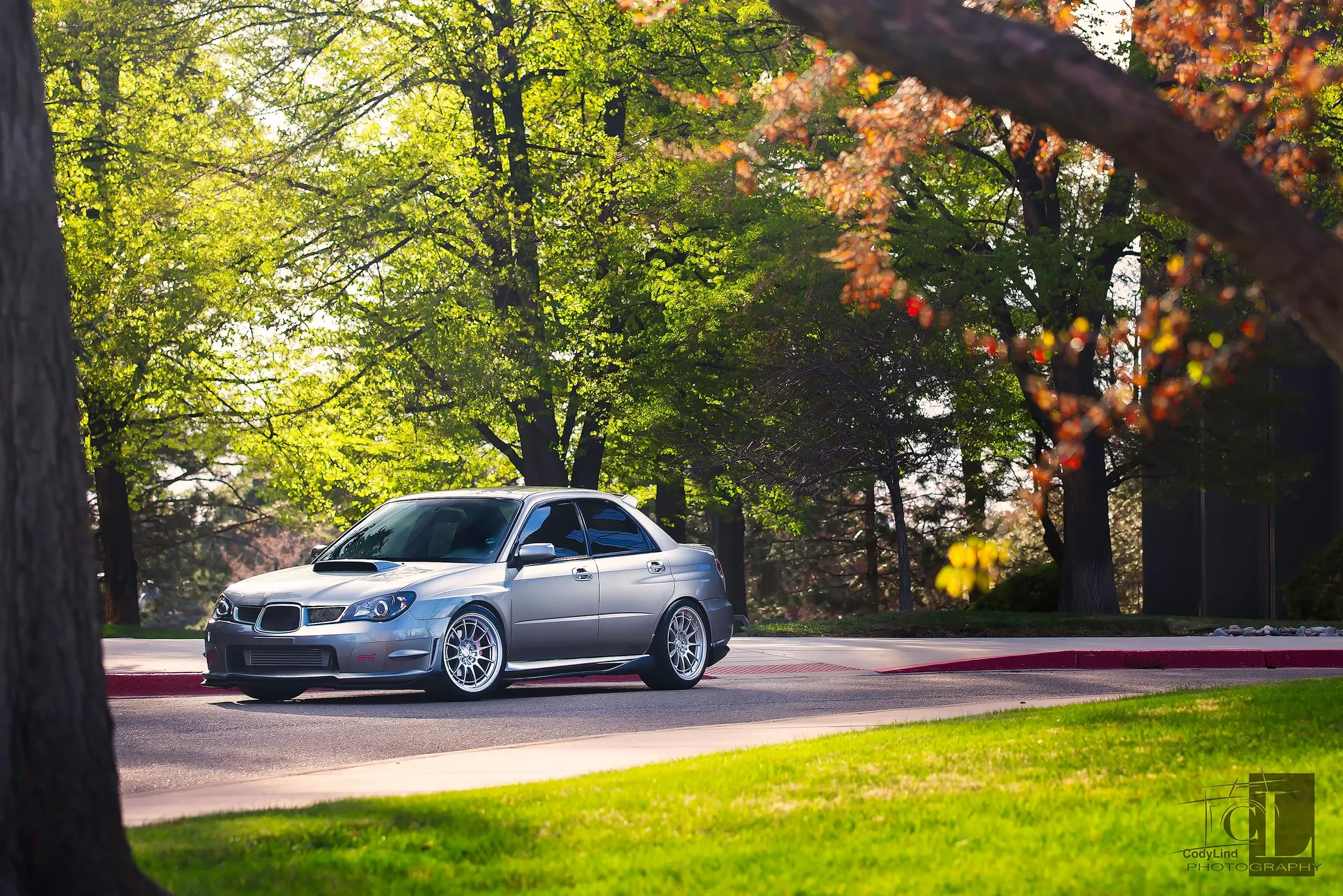 A silver Subaru Impreza WRX STI parked on a street with trees and green grass around, during daytime in spring with sunlight filtering through leaves.