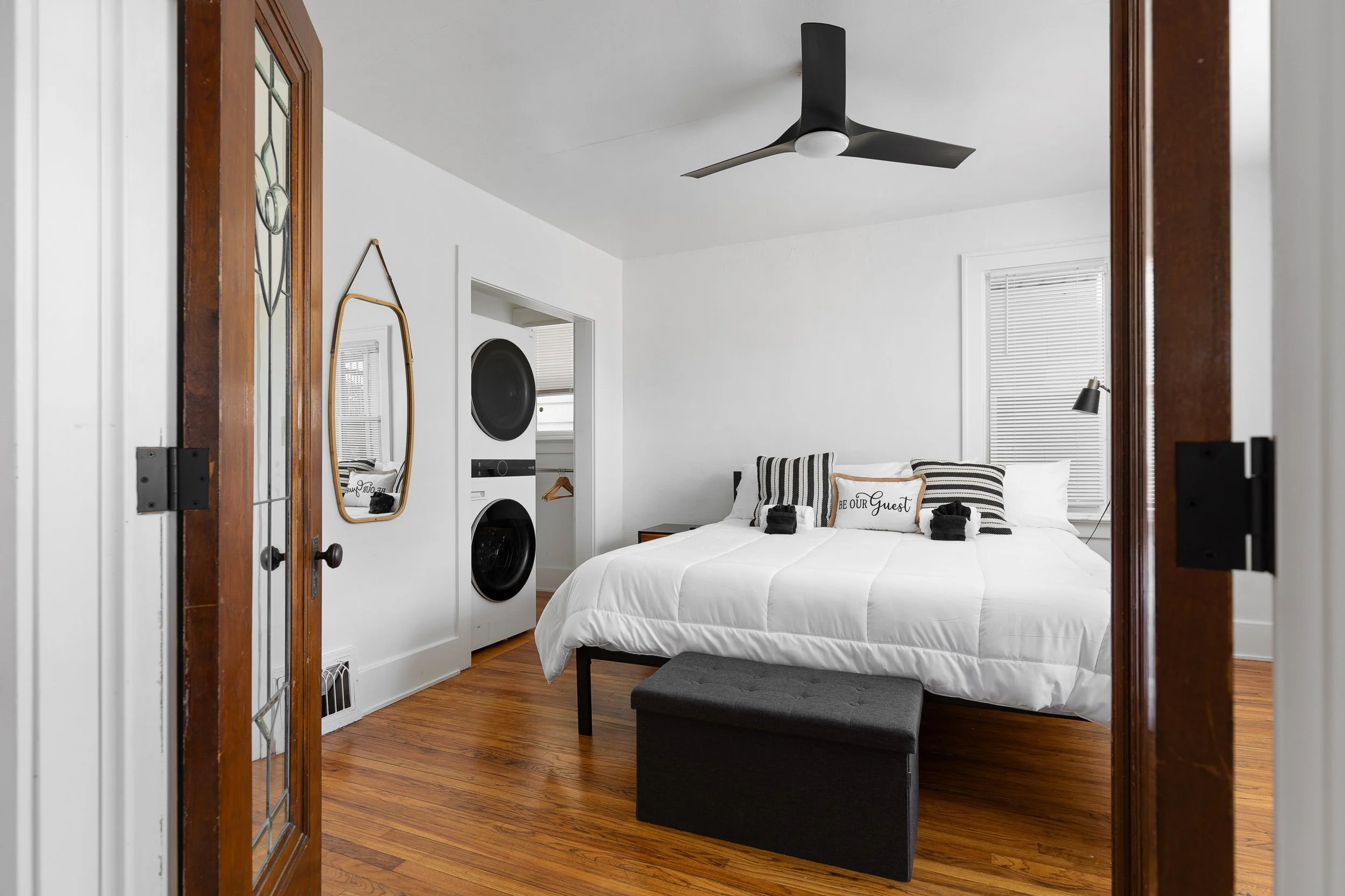 Bedroom with white walls, hardwood floors, a bed with white bedding, black and white pillows, and a black ottoman at the foot of the bed. There is a ceiling fan, a window with blinds, a black wall-mounted lamp, and a laundry area with stacked washer 