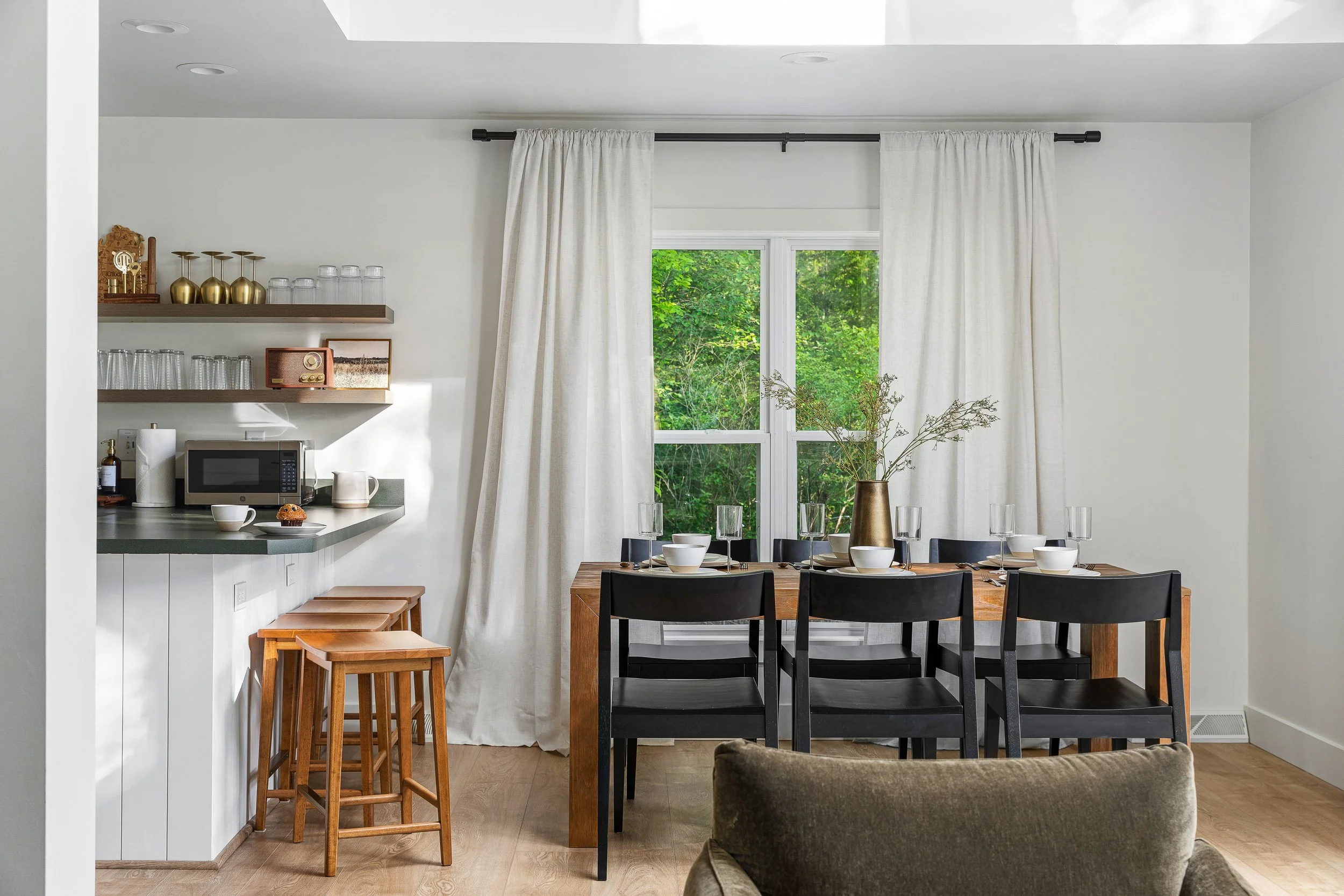 Dining area with a wooden table, black chairs, a vase with branches, and a window with green trees outside. Kitchen counter with stools, microwave, and shelves with glasses and gold vases.