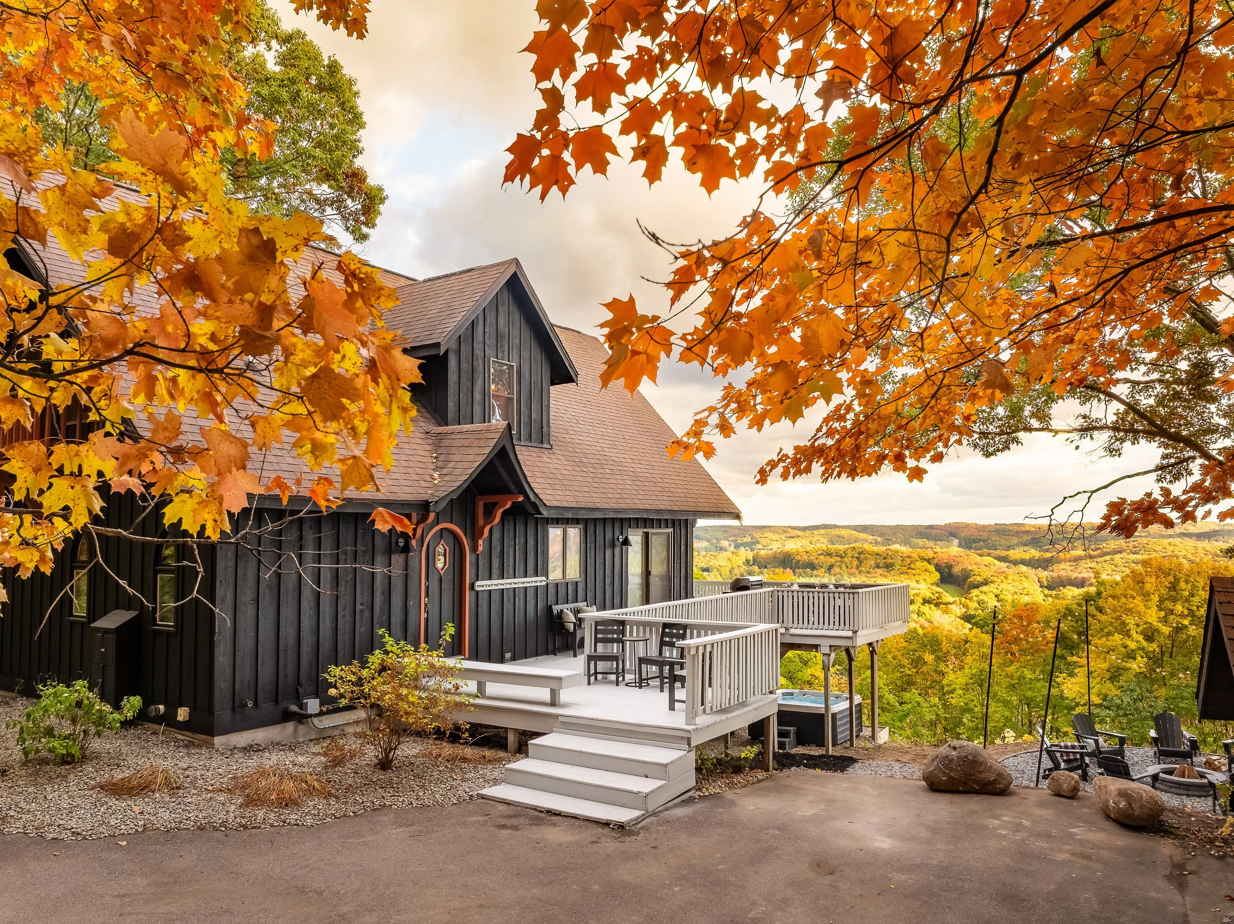 Black wooden house with a brown gabled roof and a white deck, surrounded by autumn orange and yellow leaves, overlooking a scenic landscape of rolling hills and trees in fall