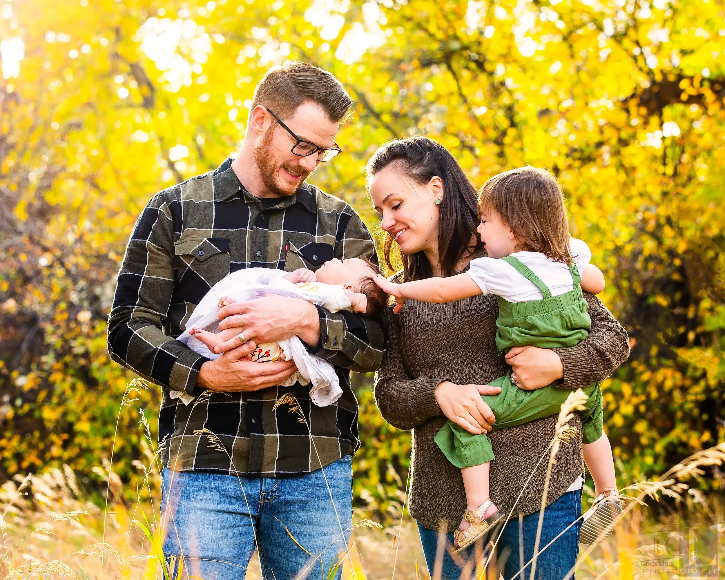 A family of four outdoors in a fall setting with yellow leaves. The father holds a newborn baby, the mother holds a young girl, and they are all smiling and touching the baby.