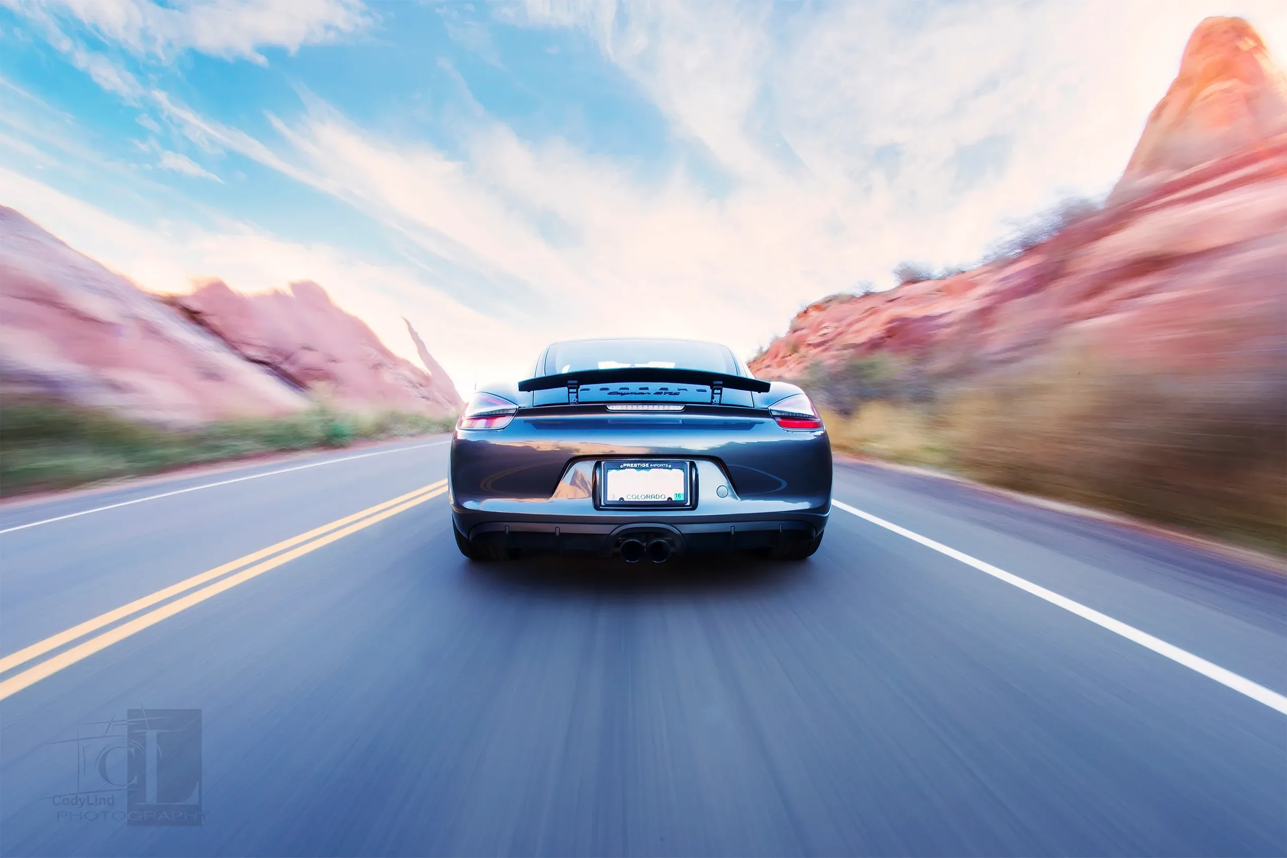 Black sports car driving on a two-lane road with red rock formations and a cloudy sky in the background.
