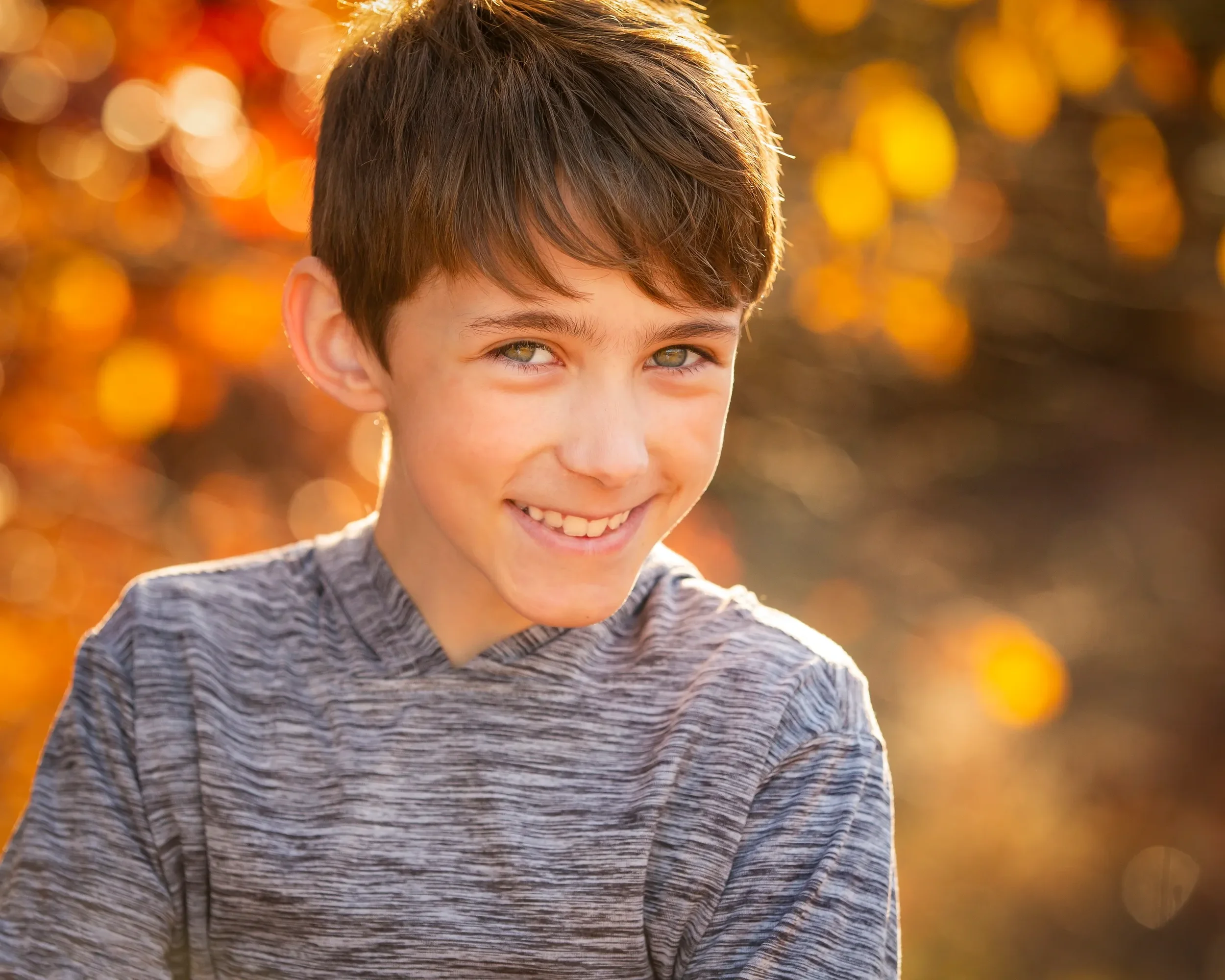 A young boy with brown hair and gray-green eyes smiling outdoors in autumn.