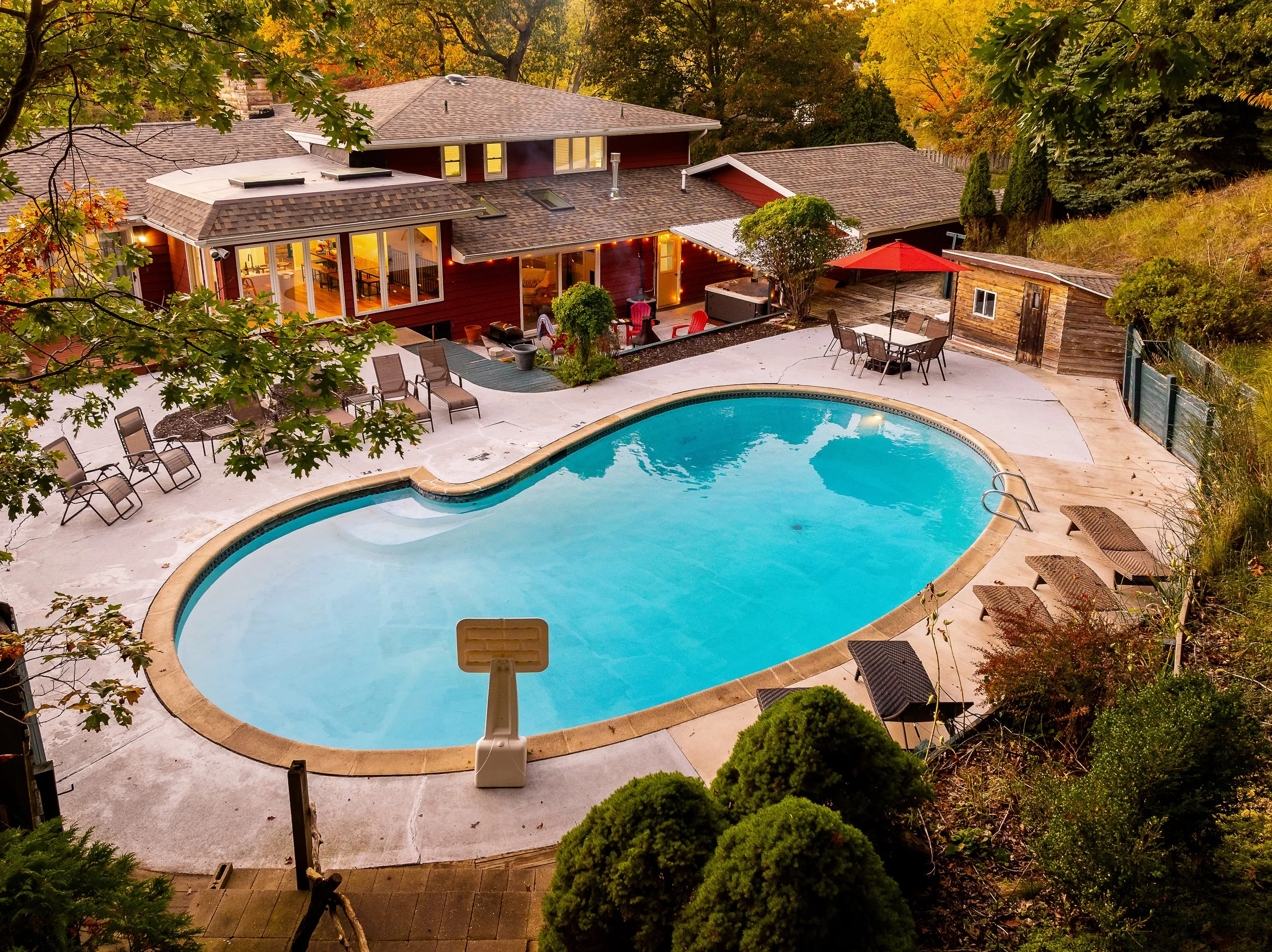 A backyard with an in-ground swimming pool surrounded by a concrete deck, outdoor patio furniture, a red umbrella, and a wooden shed, with a red house and trees in the background during autumn.