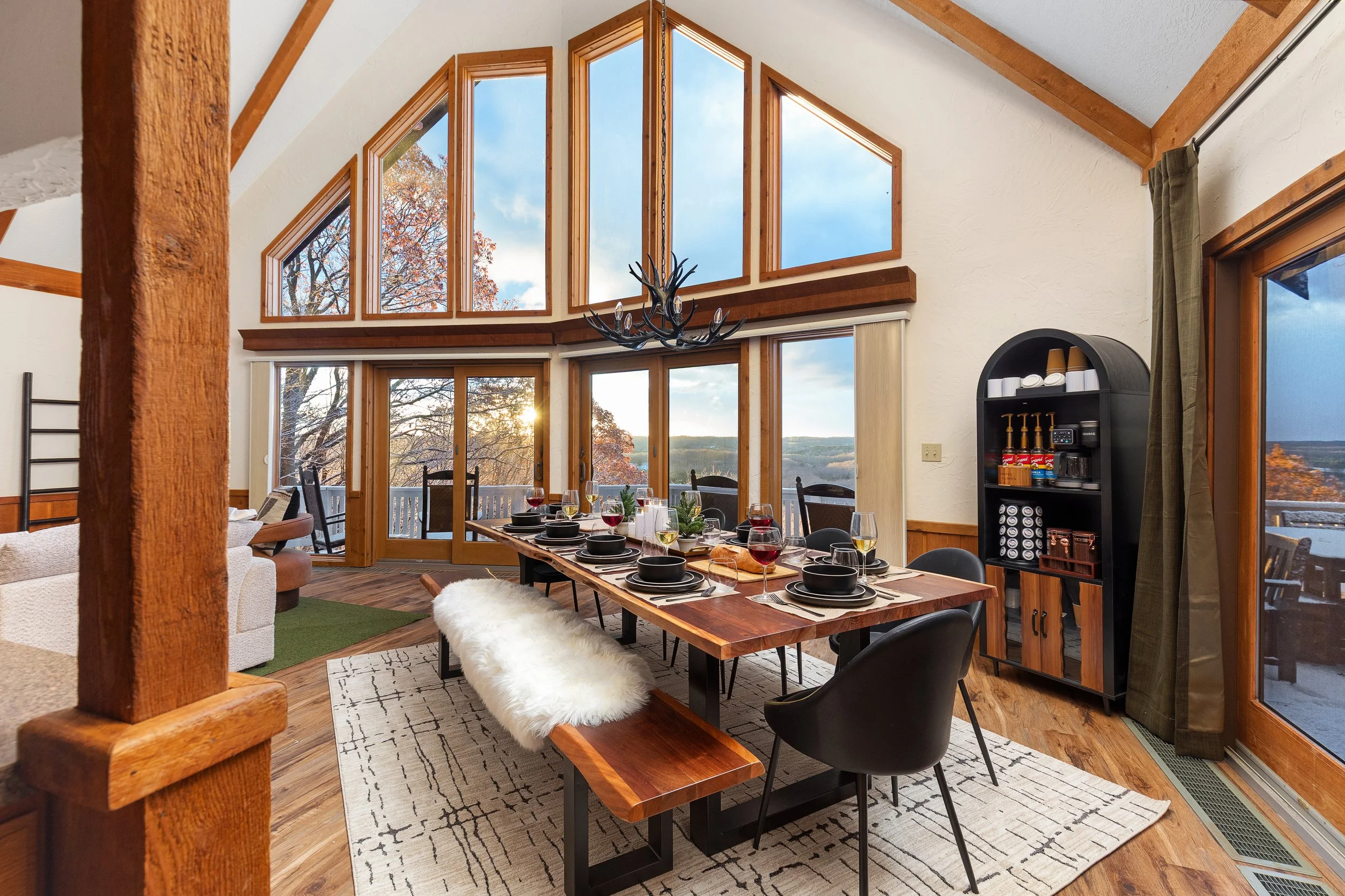 Dining room with large windows and a view of trees and water, set for a meal with black plates, wine glasses, and candles, with a rustic chandelier overhead.