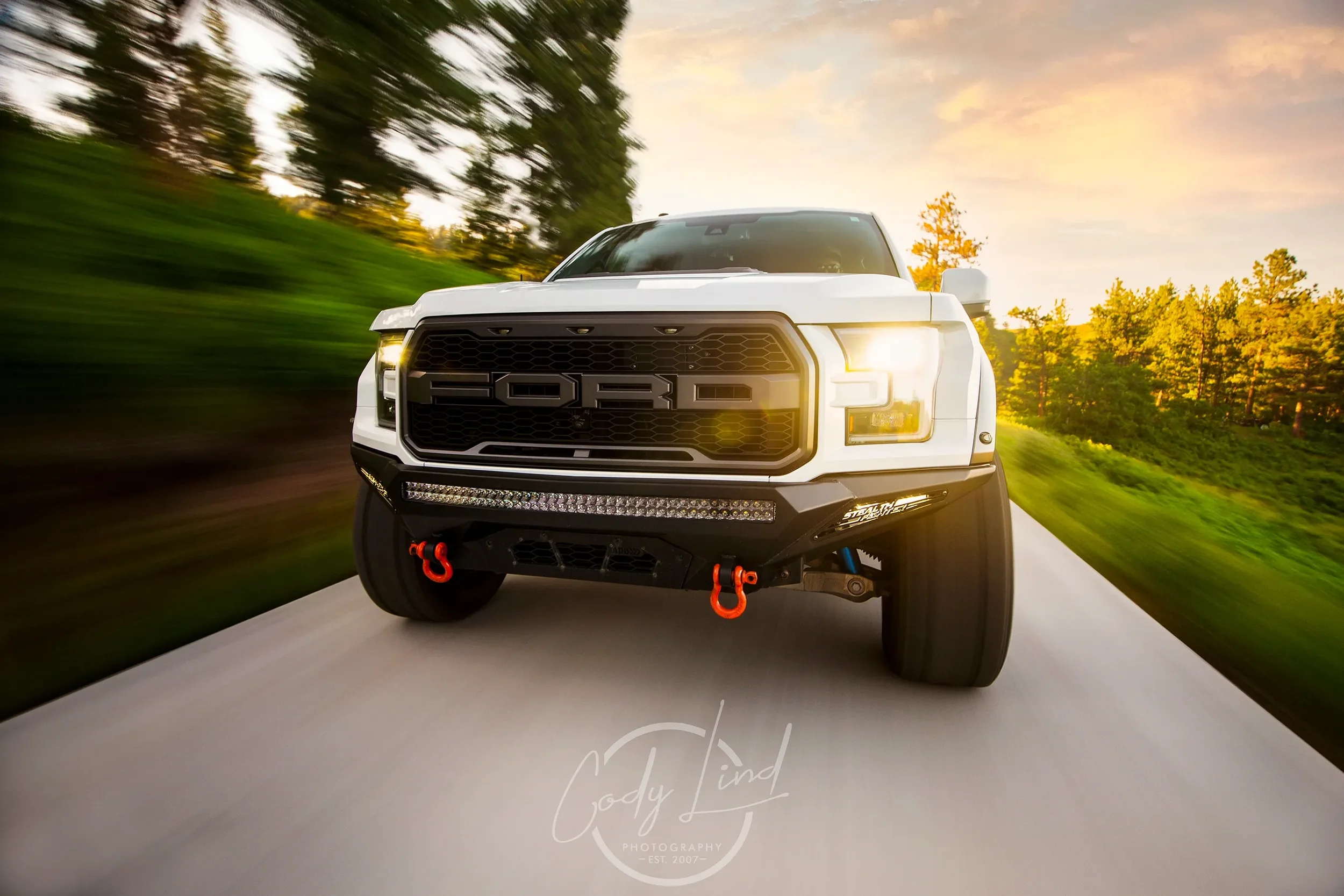 A white Ford pickup truck driving on a road surrounded by greenery with trees and a sunset sky in the background.