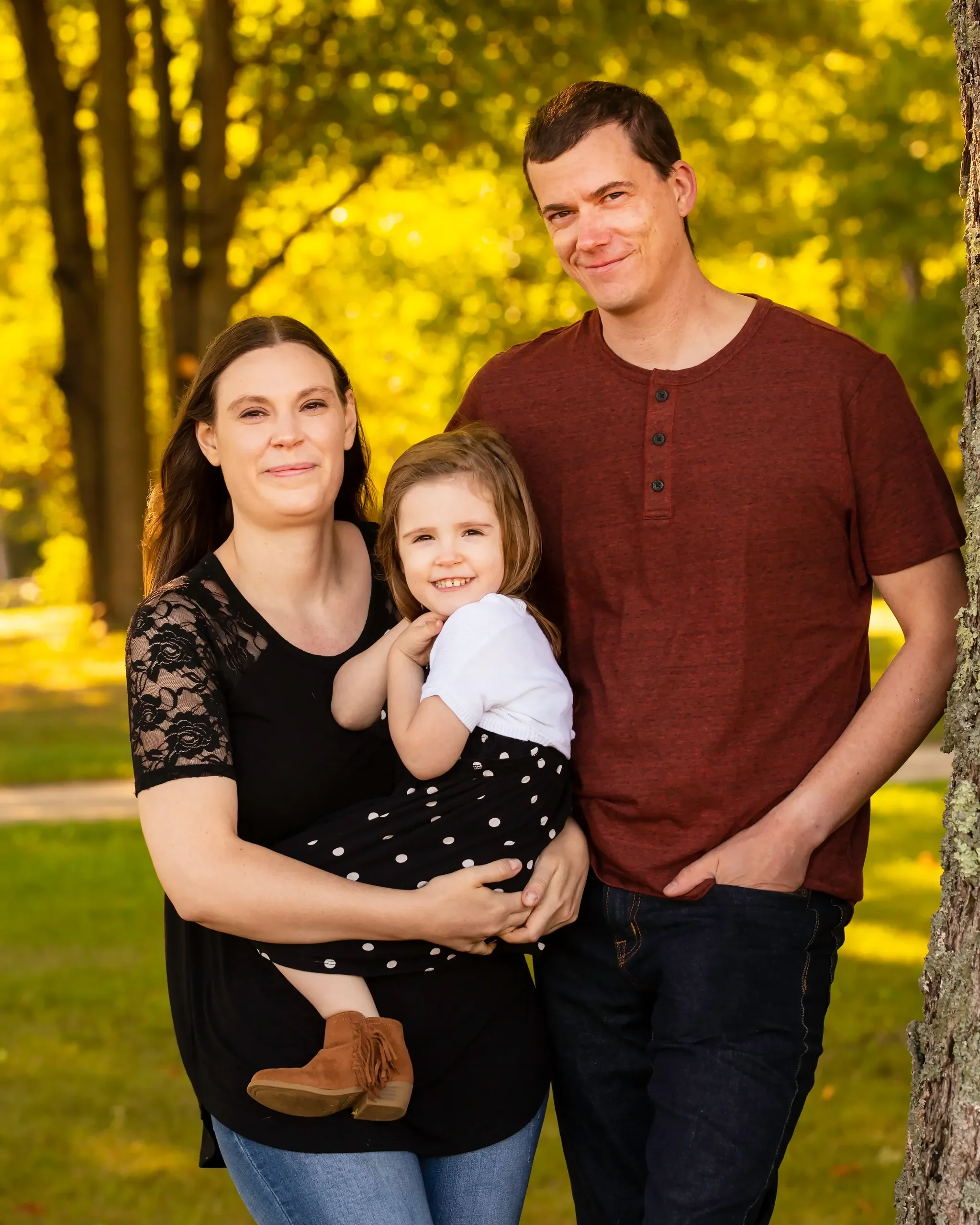Family photograph outdoors with trees and sunlight in the background, featuring a woman holding a young girl, and a man standing beside them.