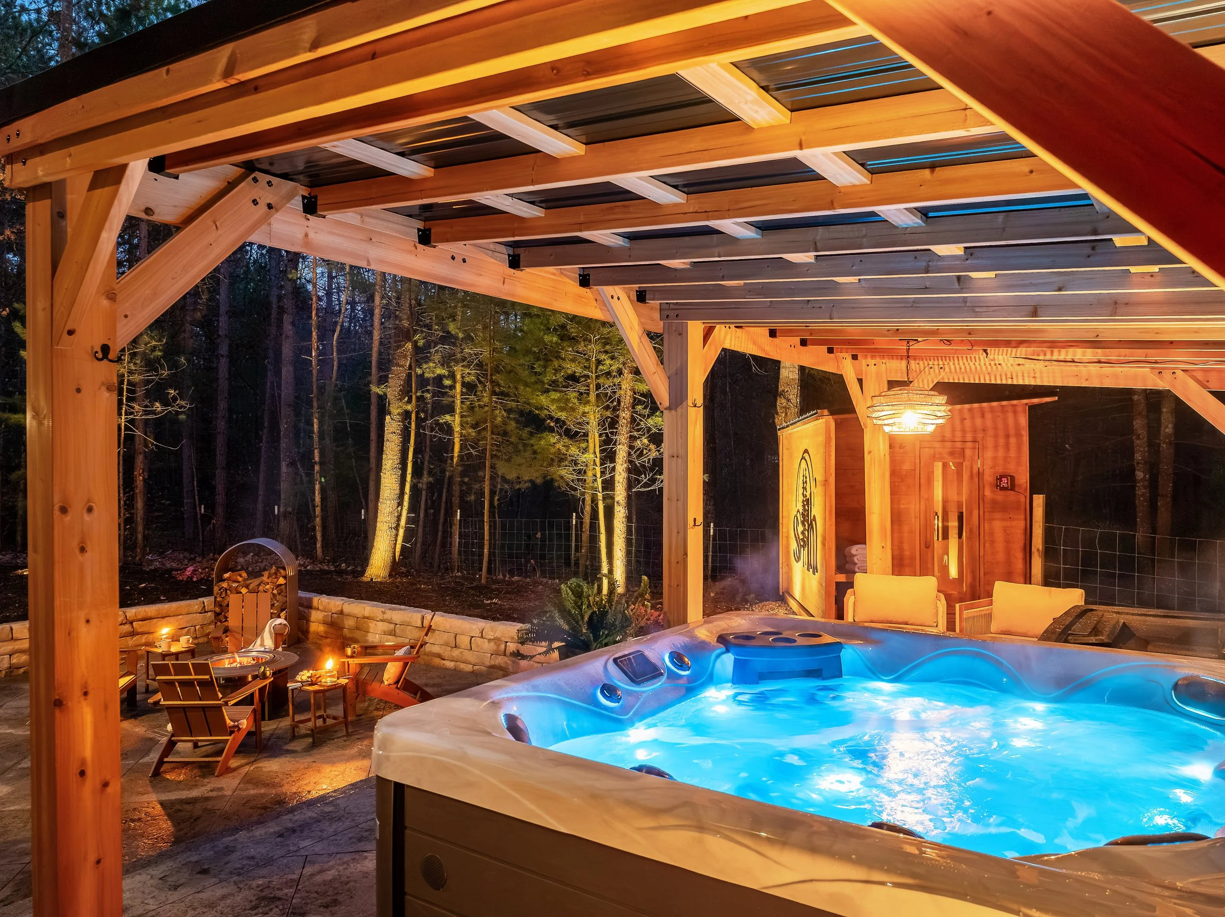 Outdoor hot tub under a wooden patio cover with string lights, surrounded by chairs and a fire pit area, in a wooded backyard at night.