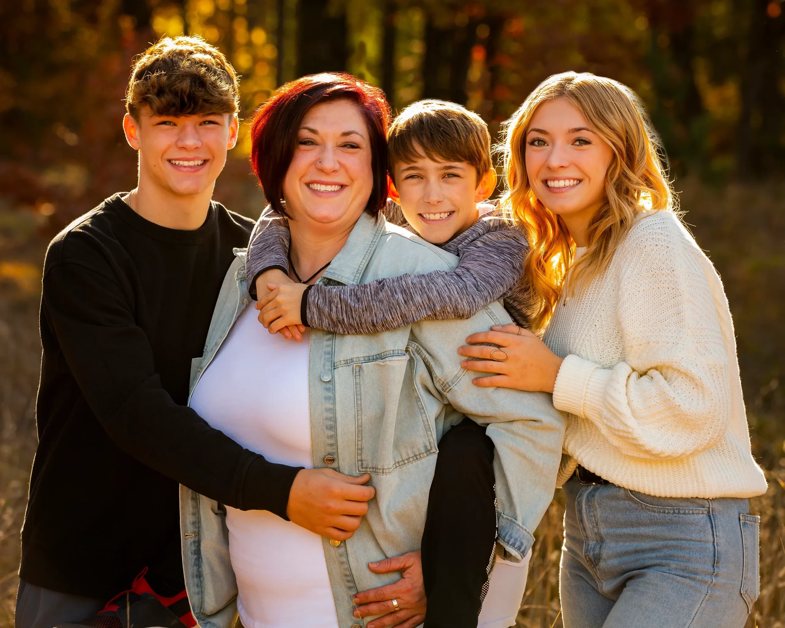 A smiling group of four people with a woman and three children outdoors during autumn, with colorful trees in the background.