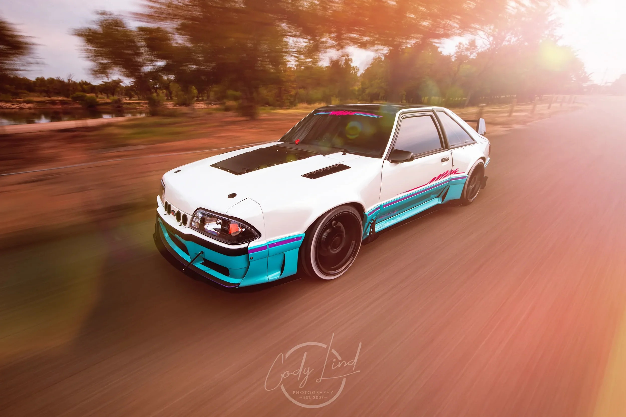 A white Japanese sports car with turquoise and pink decals racing down a dirt road during sunset.