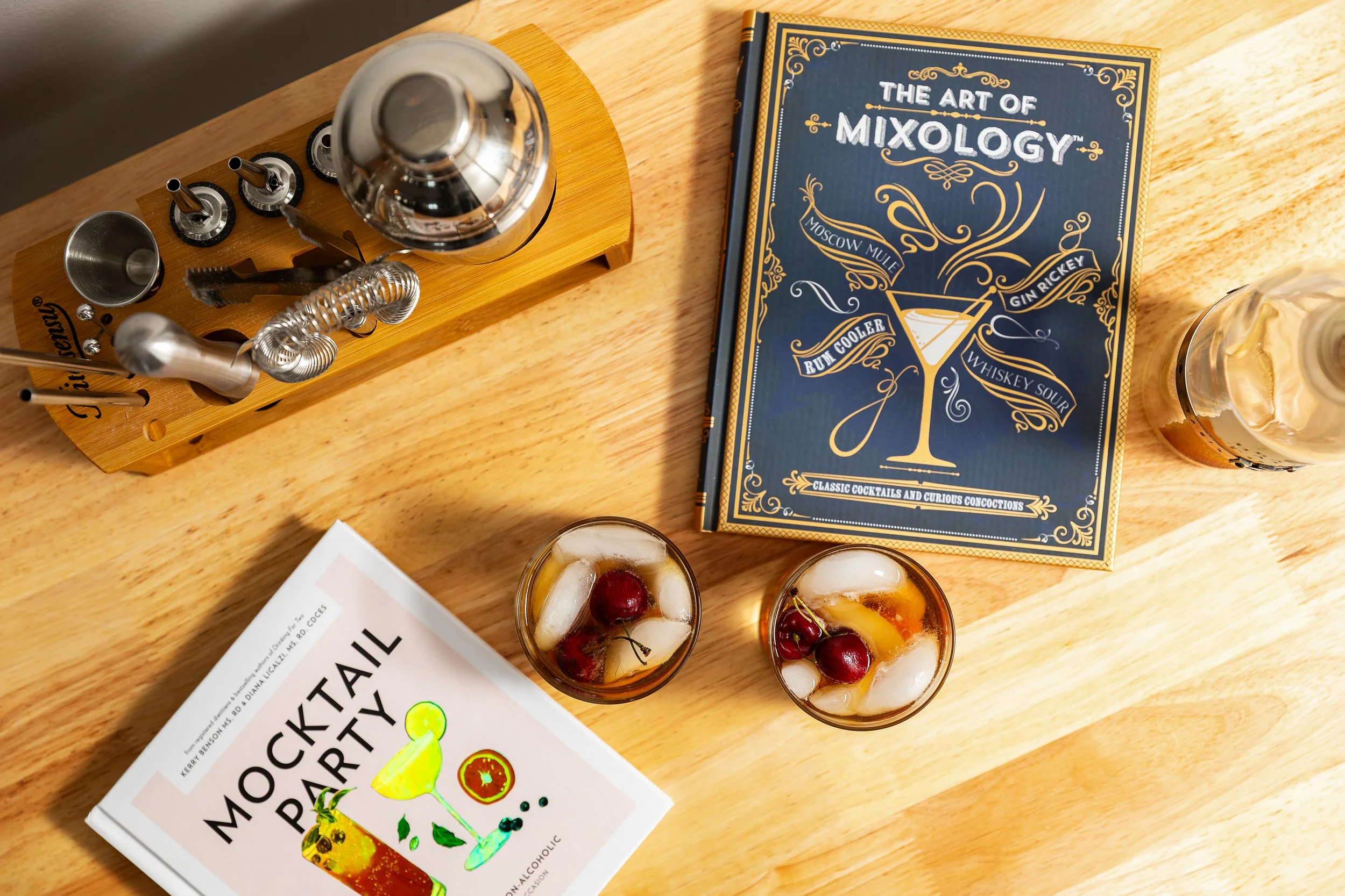 Two cocktails with ice and cherries on a wooden table, a cocktail recipe book titled 'The Art of Mixology,' a bottle of bitters, and a mockingtail party booklet.