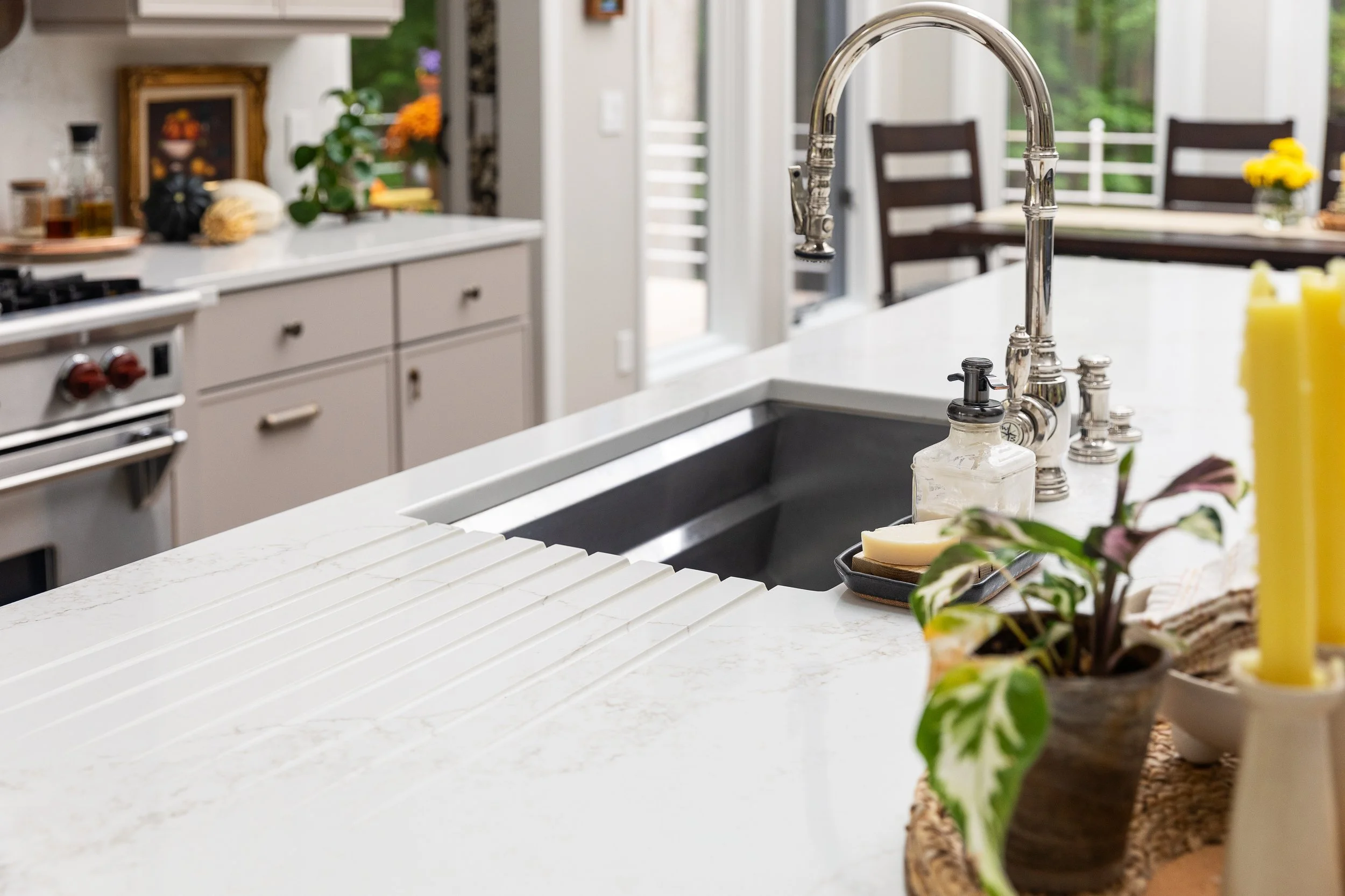 Modern kitchen with a white marble island, stainless steel sink, and chrome faucet. Decorative items and flowers on the countertop, with a view of a green outdoor area through glass doors.