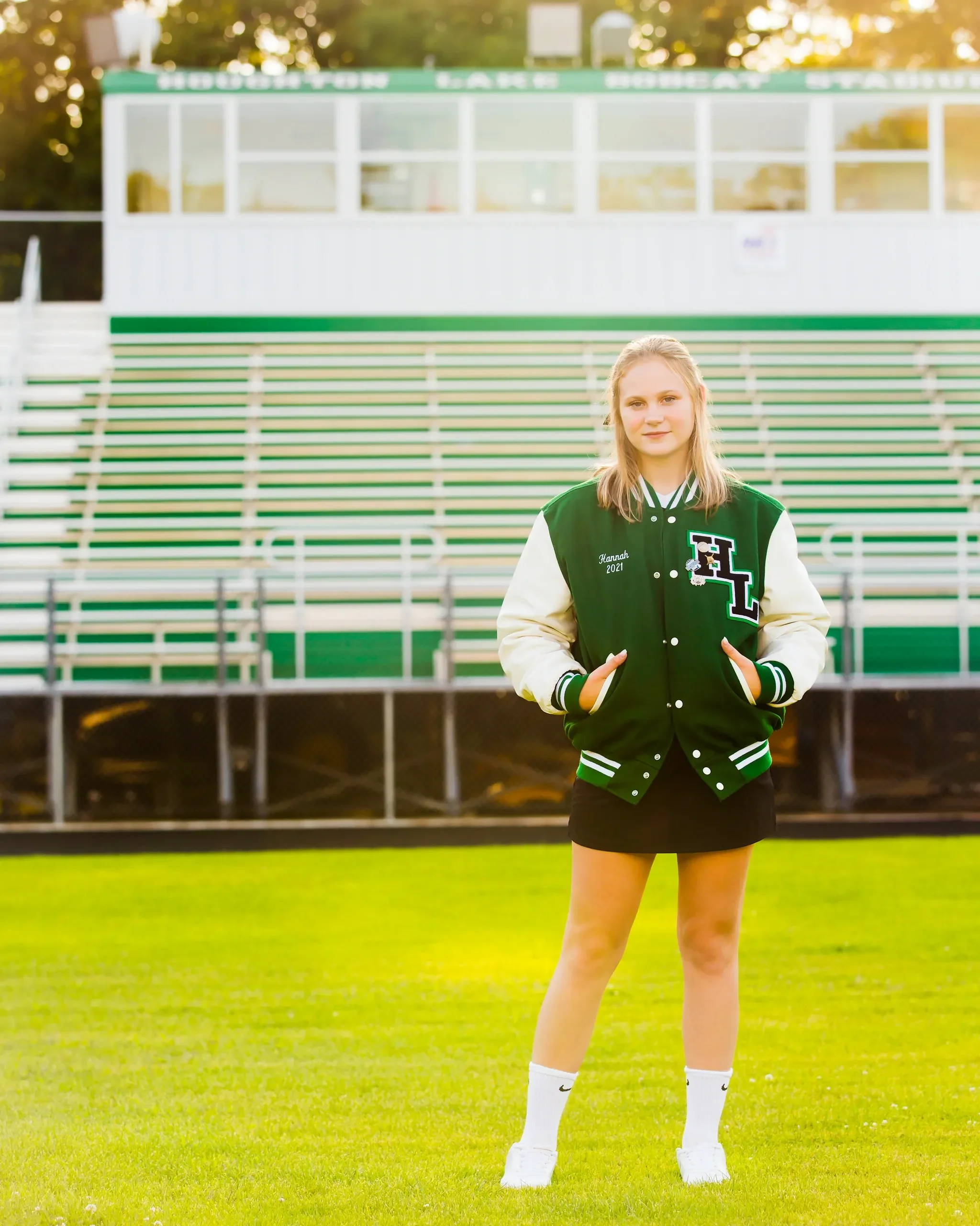 A young woman standing on a sports field with green bleachers behind her. She is wearing a green and white letterman jacket with 'Hannah 2021' embroidered on it, a black skirt, white Nike socks, and white sneakers.