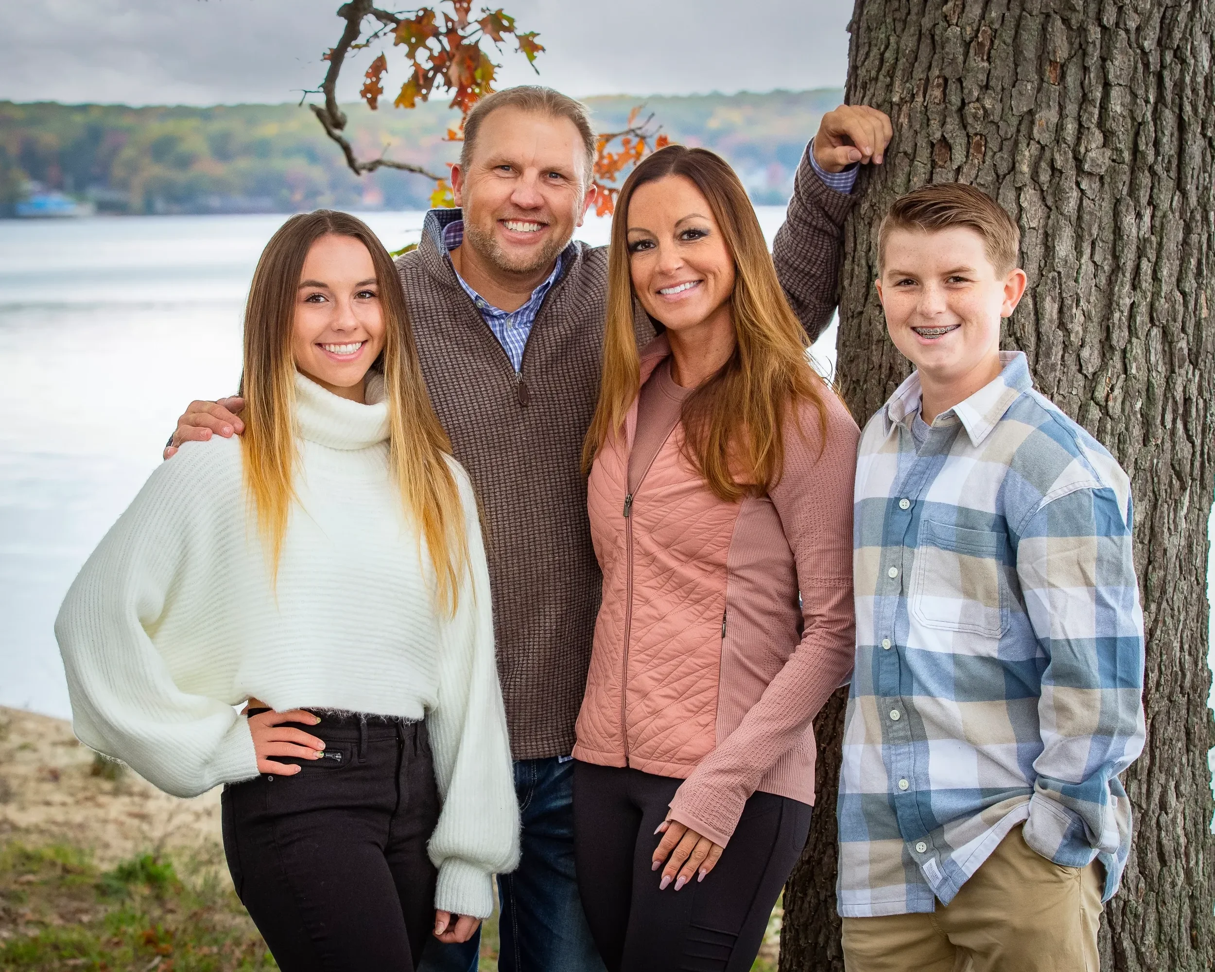 A family of five standing outdoors near a lake with trees in the background. The family members are smiling and standing close together. A woman is wearing a white turtleneck sweater, a man in a brown plaid shirt, a woman in a pink jacket, a teenage 