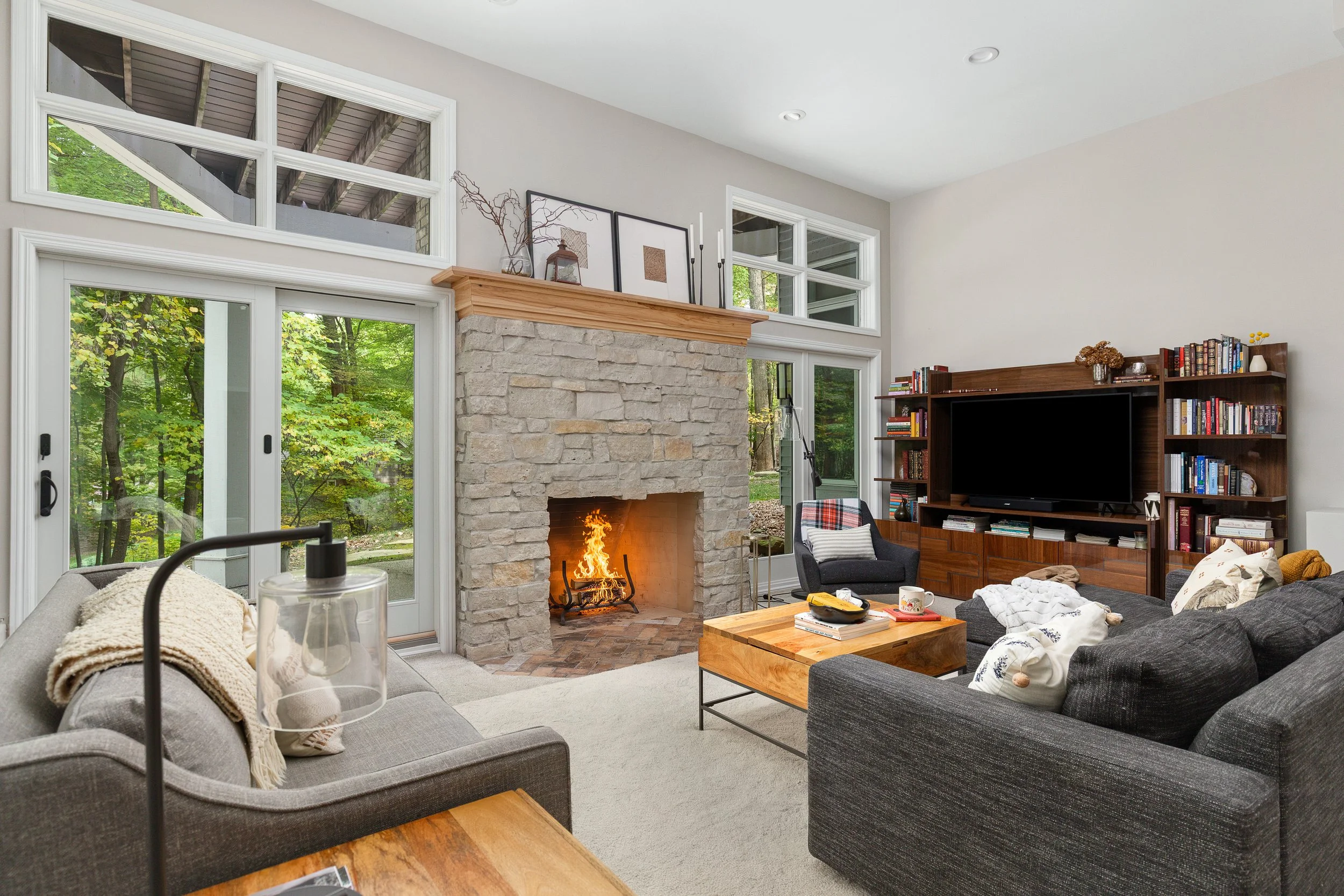 Living room with a stone fireplace, a large window and sliding glass door showing green trees outside, dark sofas with pillows, a wooden coffee table, a wall-mounted TV, and a bookshelf with books and decorations.