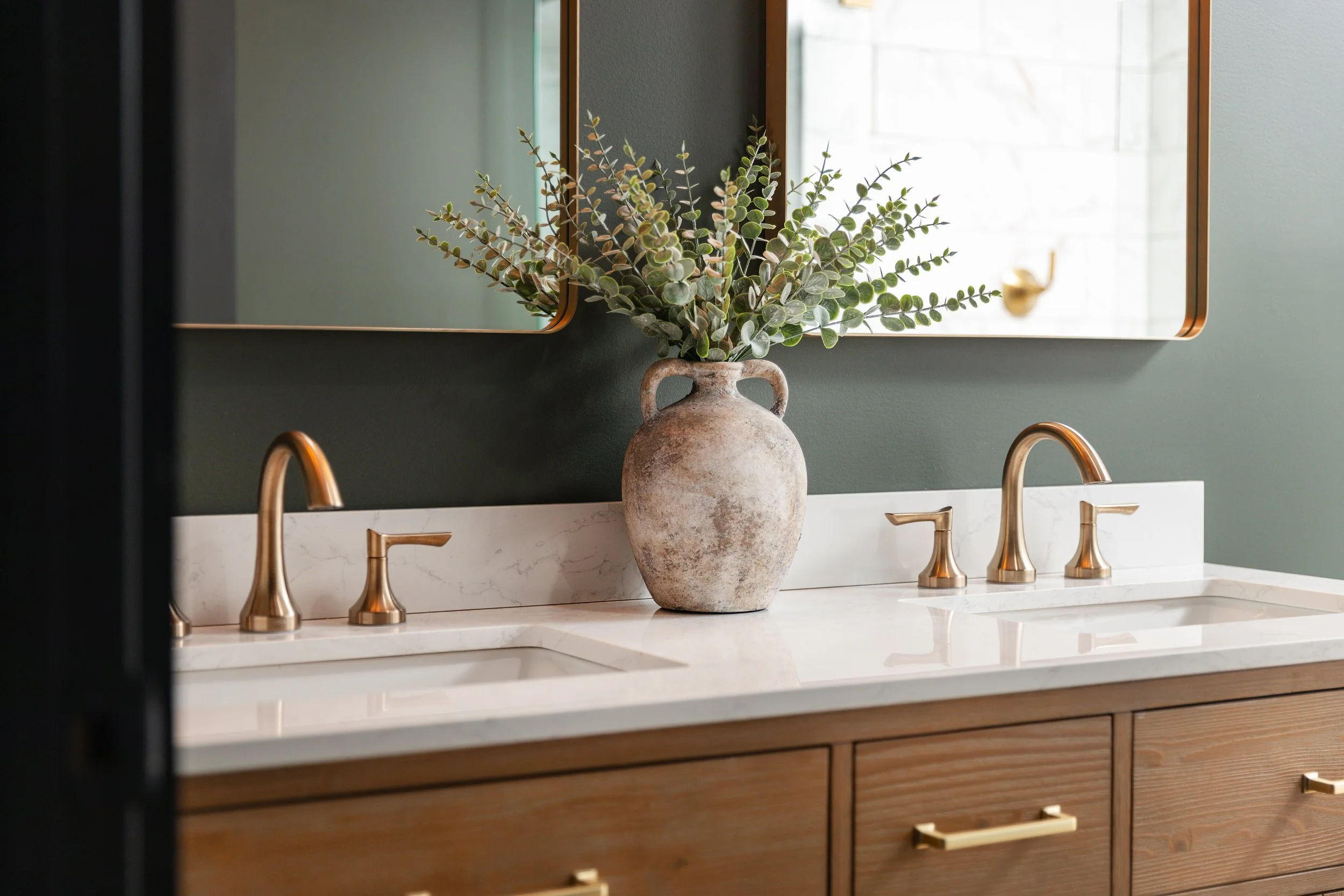 Bathroom vanity with dual sinks, gold faucets, a large rustic vase with green foliage, and a mirror on a green wall.