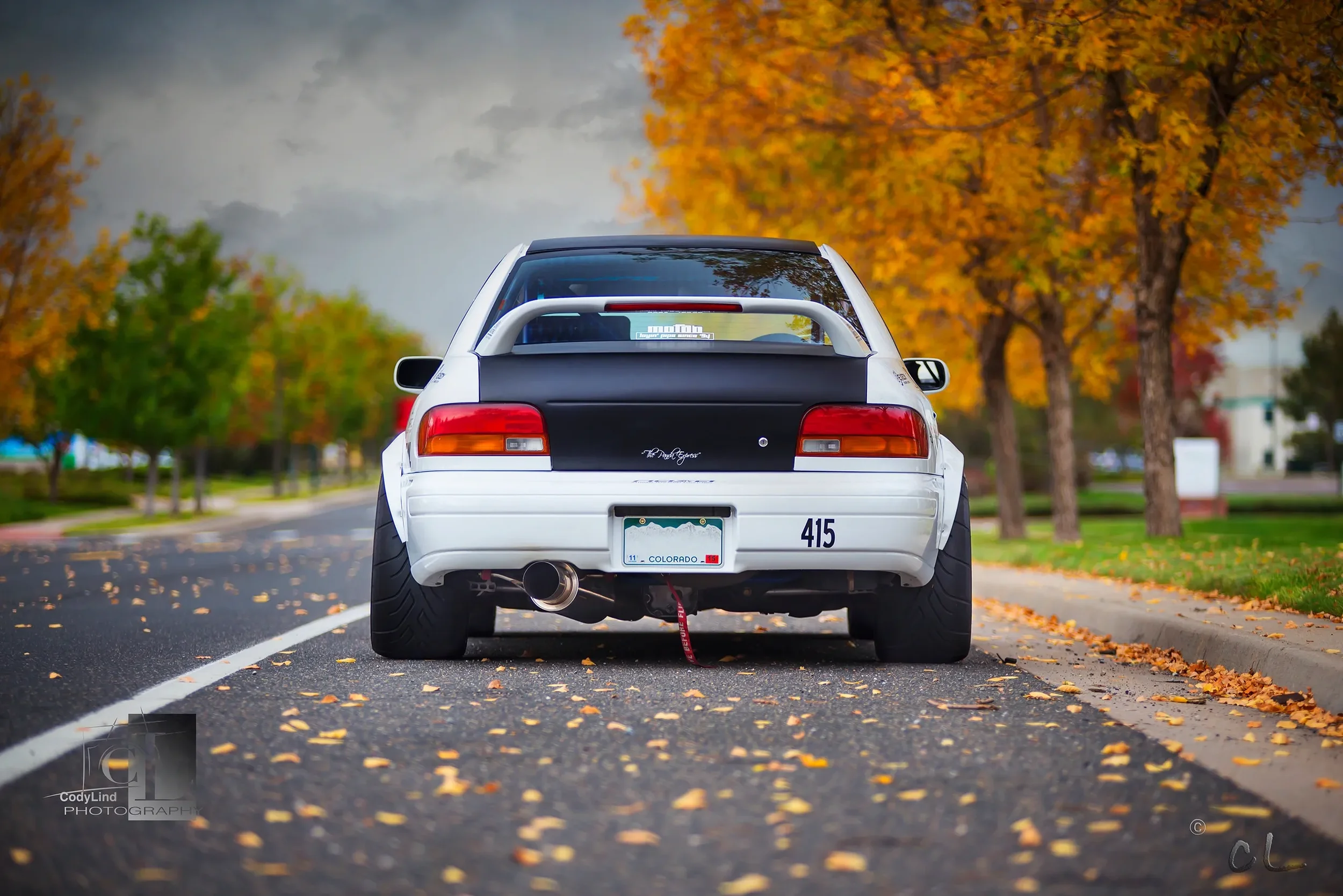 Rear view of a white sports car with wide tires parked on a street surrounded by trees with autumn-colored leaves.