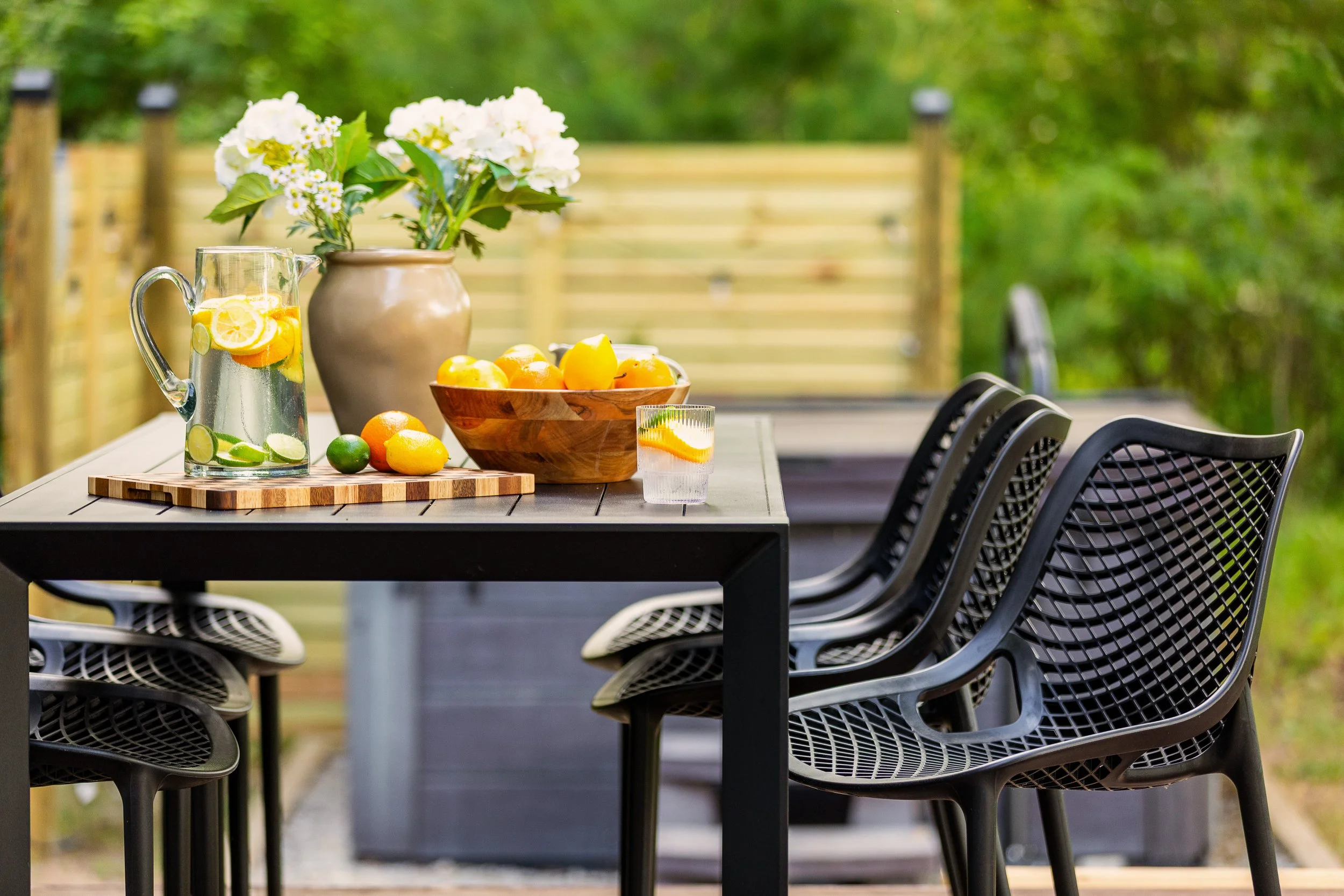 Outdoor dining table with a pitcher of lemon and cucumber infused water, a wooden bowl of citrus fruits, a glass of water with lemon, and white flowers in a beige vase, set against a wooden fence and green trees.