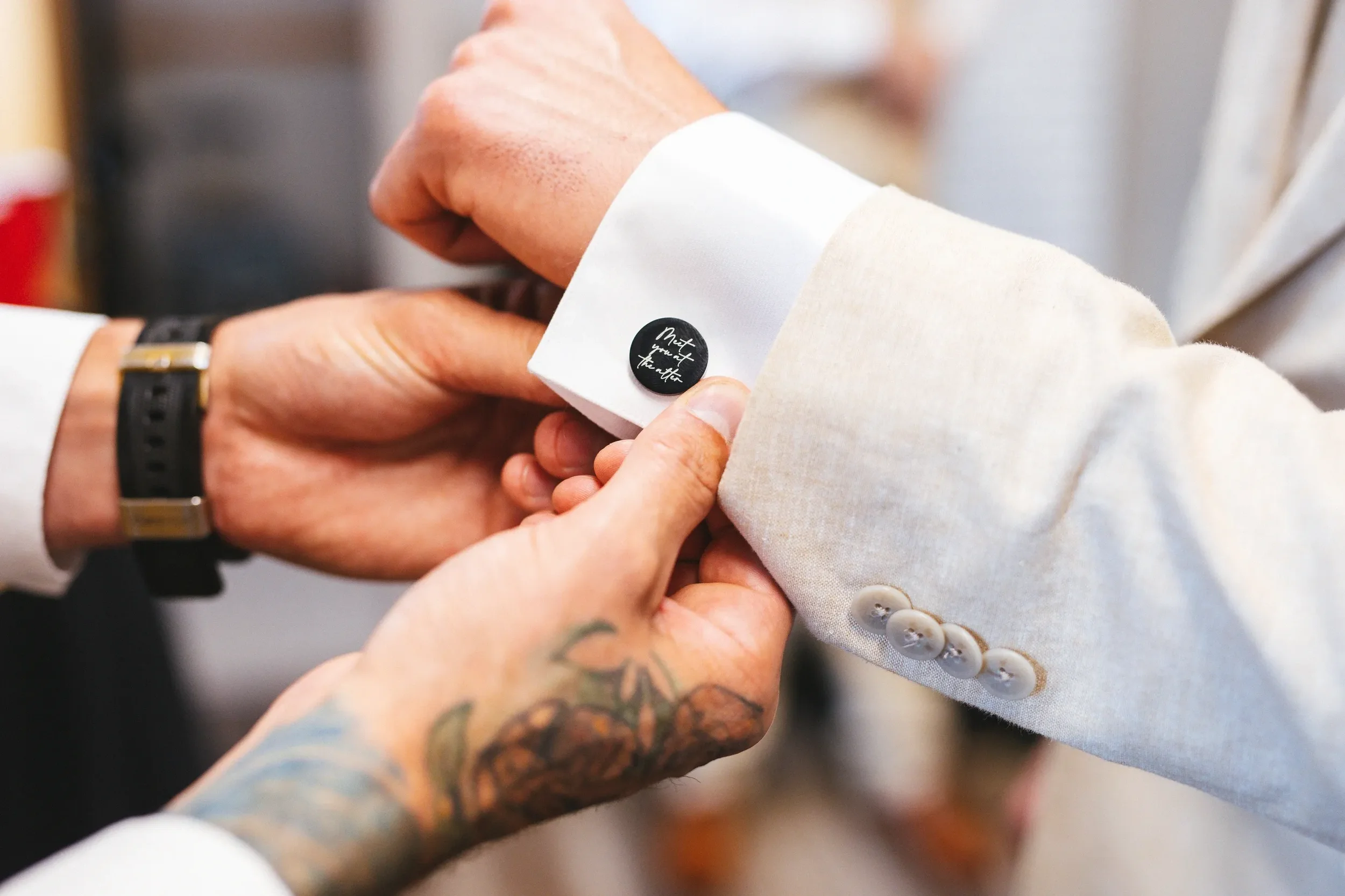 Close-up of two people adjusting the cuffs of a light-colored suit jacket with a black cufflink that says 'Made for each other.' One person's hand has a tattoo and a black wristwatch, while the other person's hand supports the cuff.