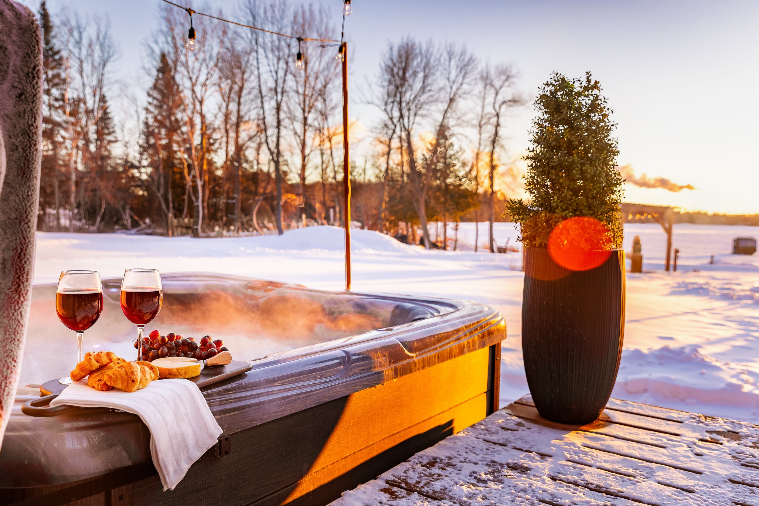 A hot tub outdoors in a snowy setting with two glasses of red wine, grapes, croissants, and cheese on a tray. There is a potted plant nearby, and string lights hang above. The background shows a snowy landscape with trees and a sunset sky.