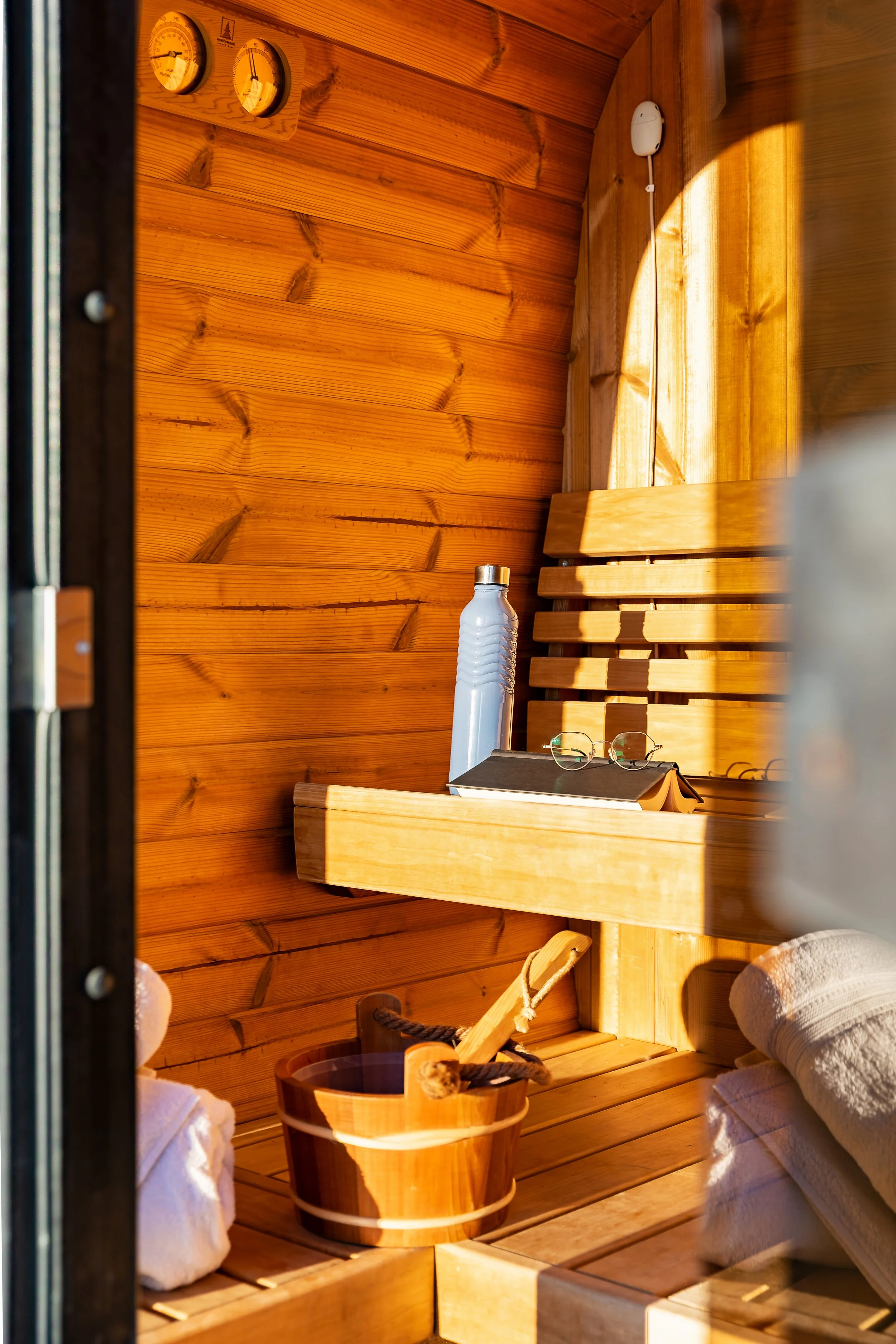 Interior of a wooden sauna with shelves, a water bottle, a pair of glasses, a thermometer and humidity gauge on the wall, towels, and a wooden bucket with a ladle.