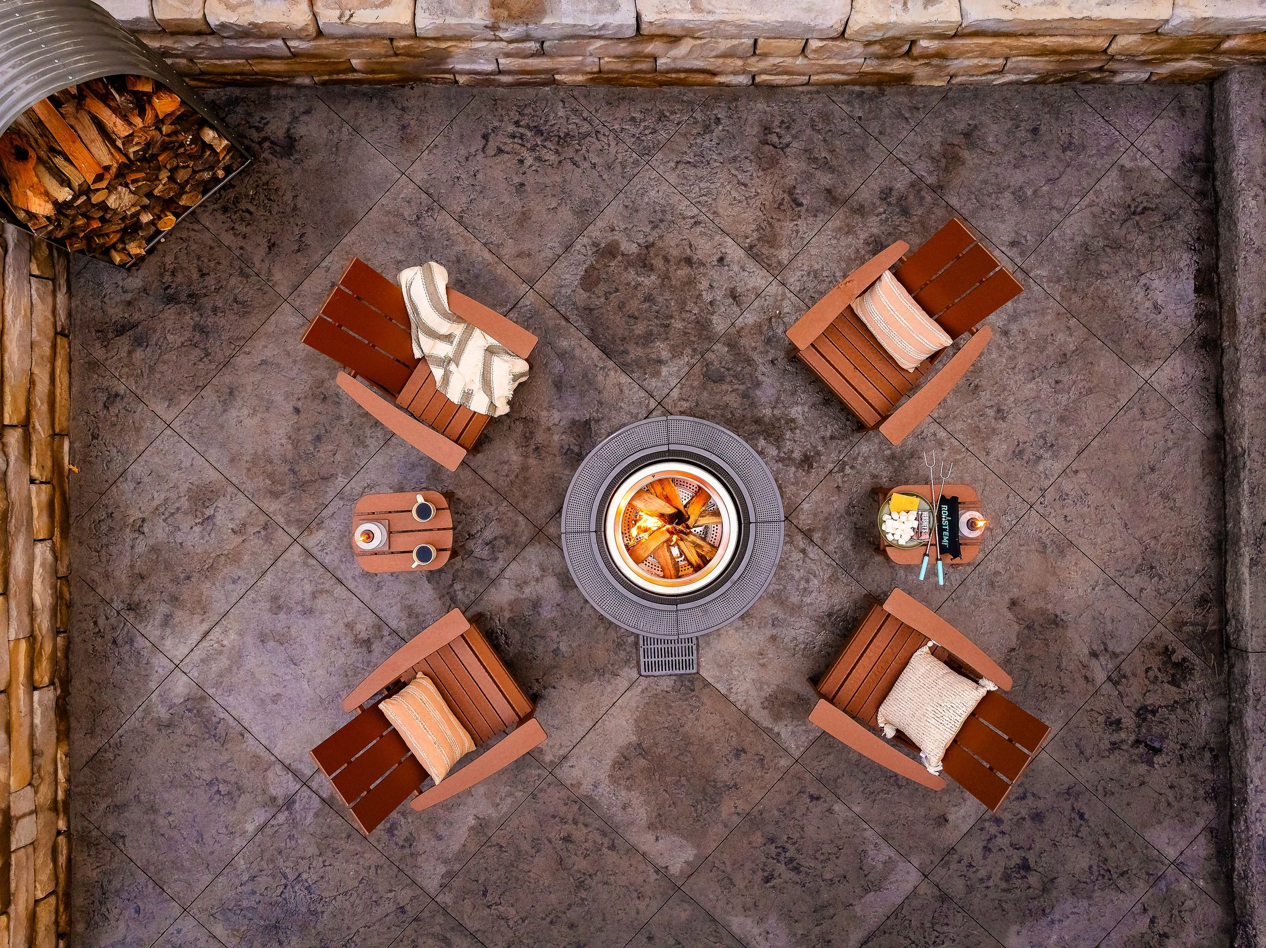 Top-down view of an outdoor seating area with four wooden chairs arranged in a square around a fire pit. Each chair has a cushion or blanket; two small tables hold beverages, snacks, and utensils. A pile of firewood is in the corner.
