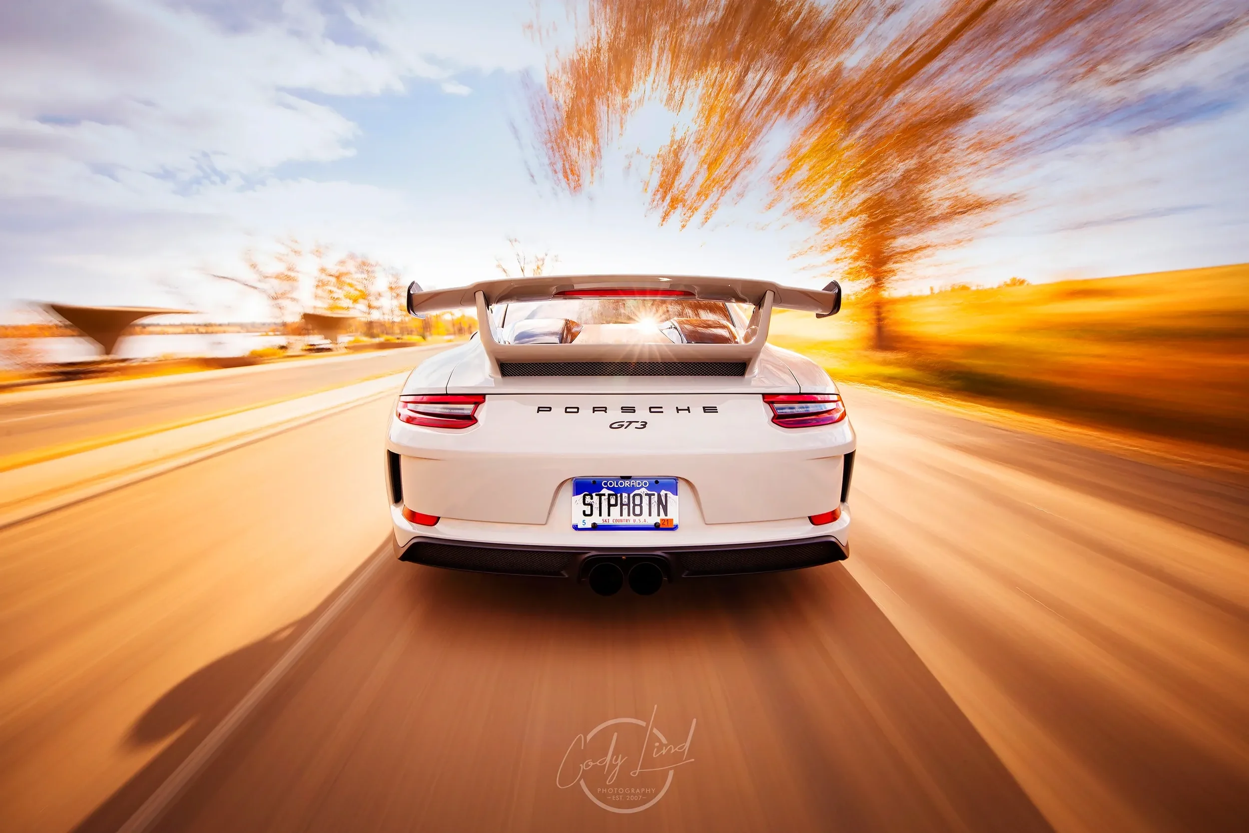 A white Porsche GT3 racing down a road with a sunset sky and autumn trees in the background.