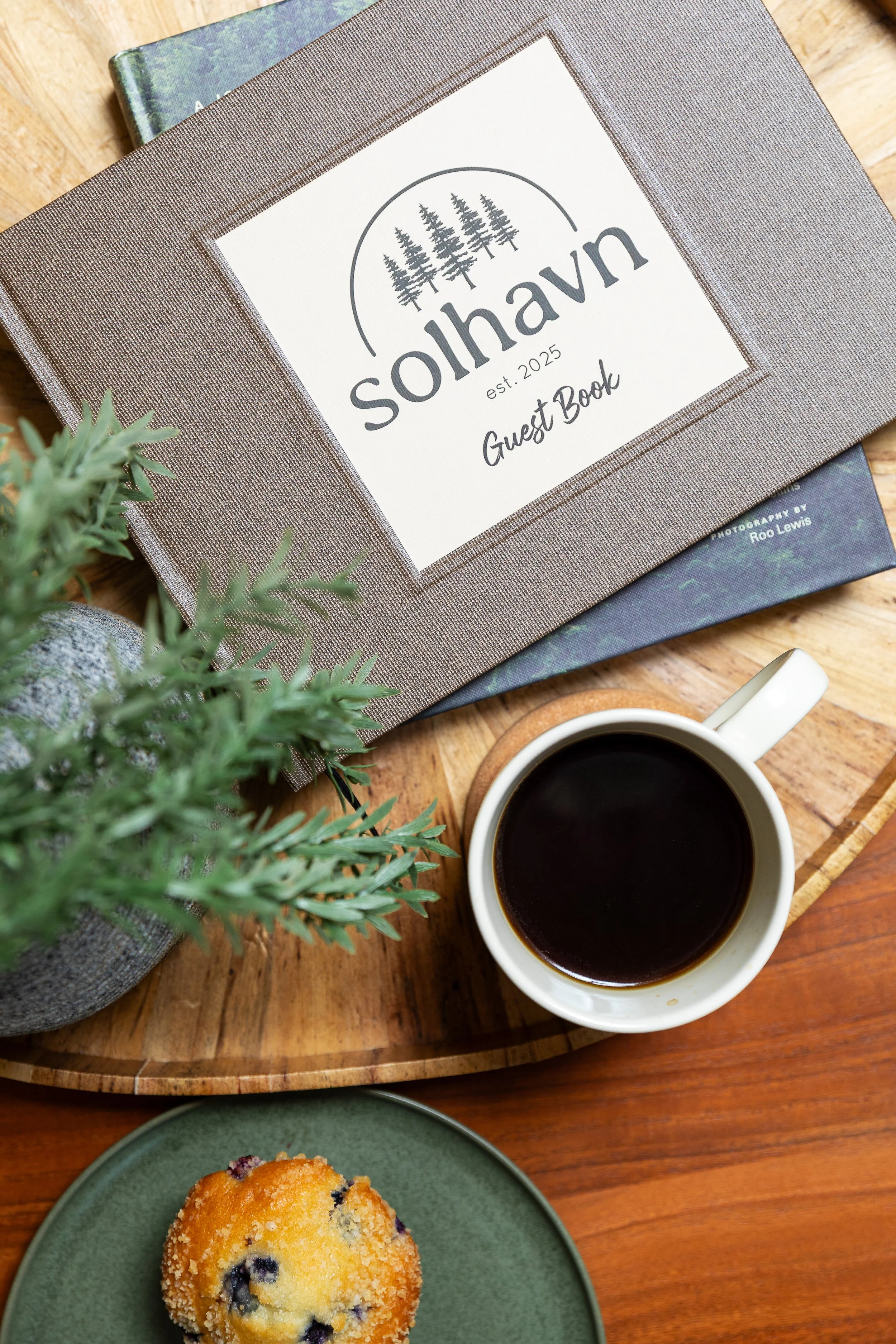 A guest book with a logo of trees and the text "solhavn est. 2025 Guest Book" sits on a wooden table. Nearby is a cup of black coffee on a coaster, a muffin on a green plate, and some green foliage.