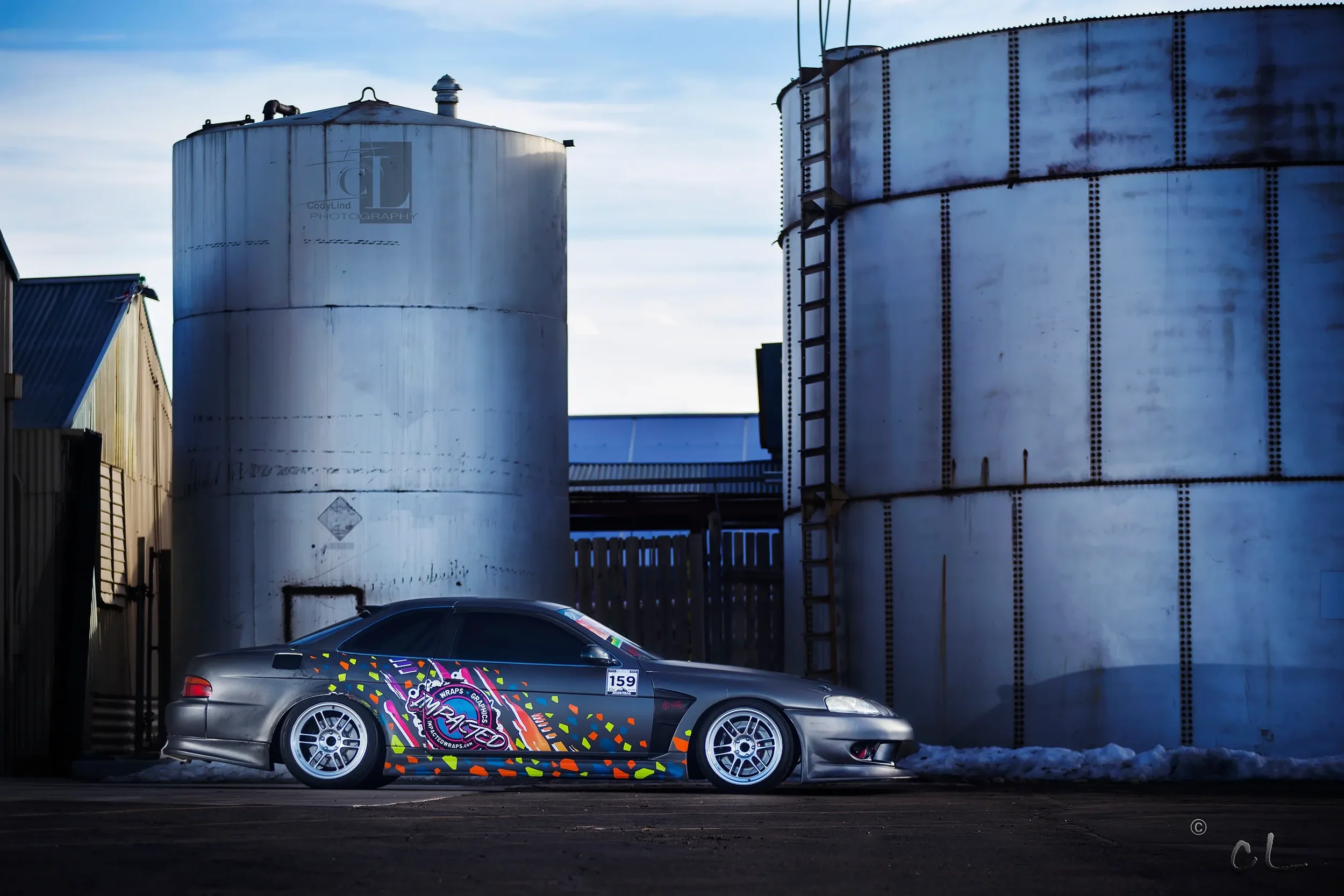A modified gray sports car with colorful graphics and the word 'Impact' on the side, parked in front of large industrial silos during twilight.