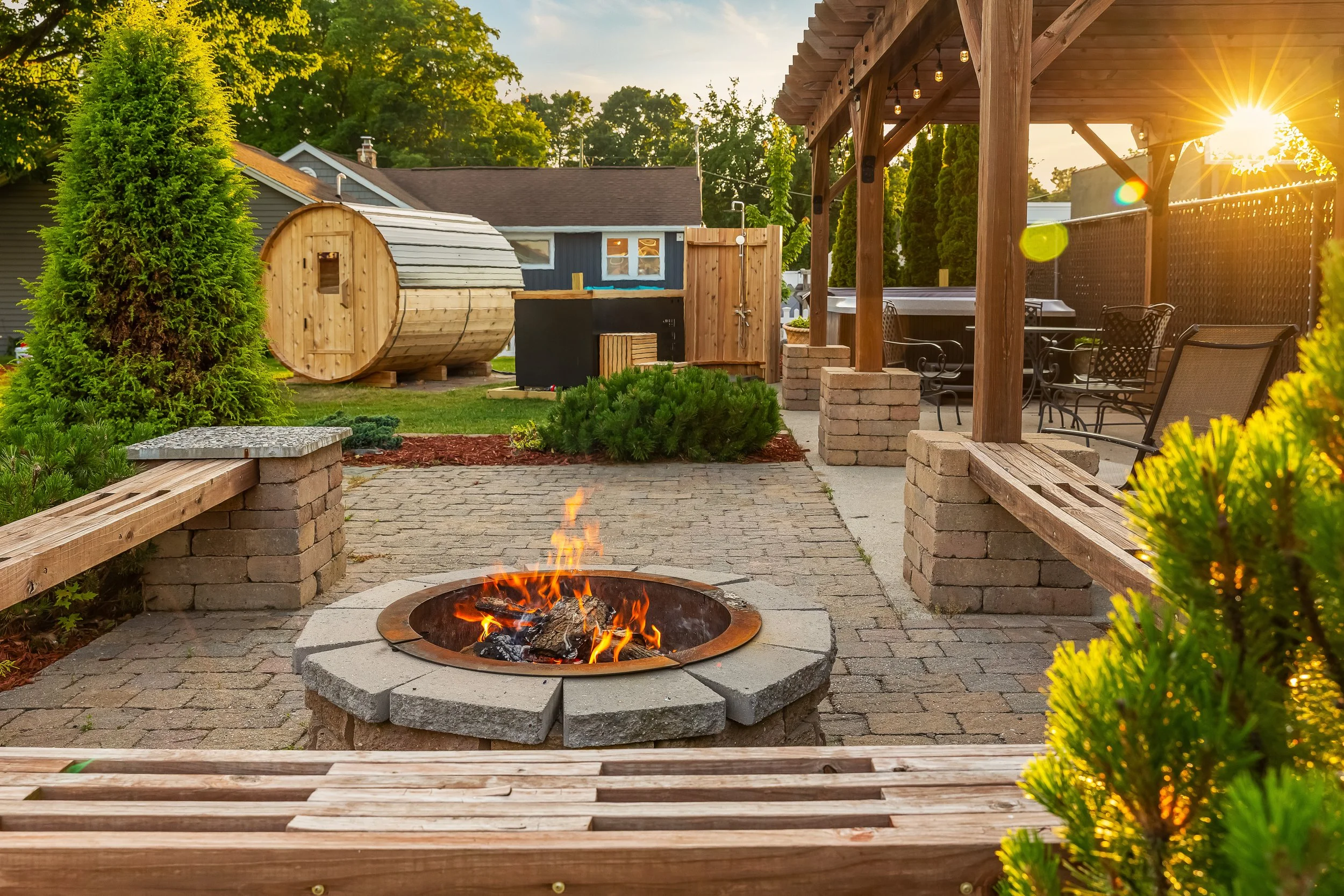 A backyard patio with a fire pit, wooden benches, and an outdoor seating area, with a small shed, trees, and a wooden barrel-shaped sauna in the background, during sunset.