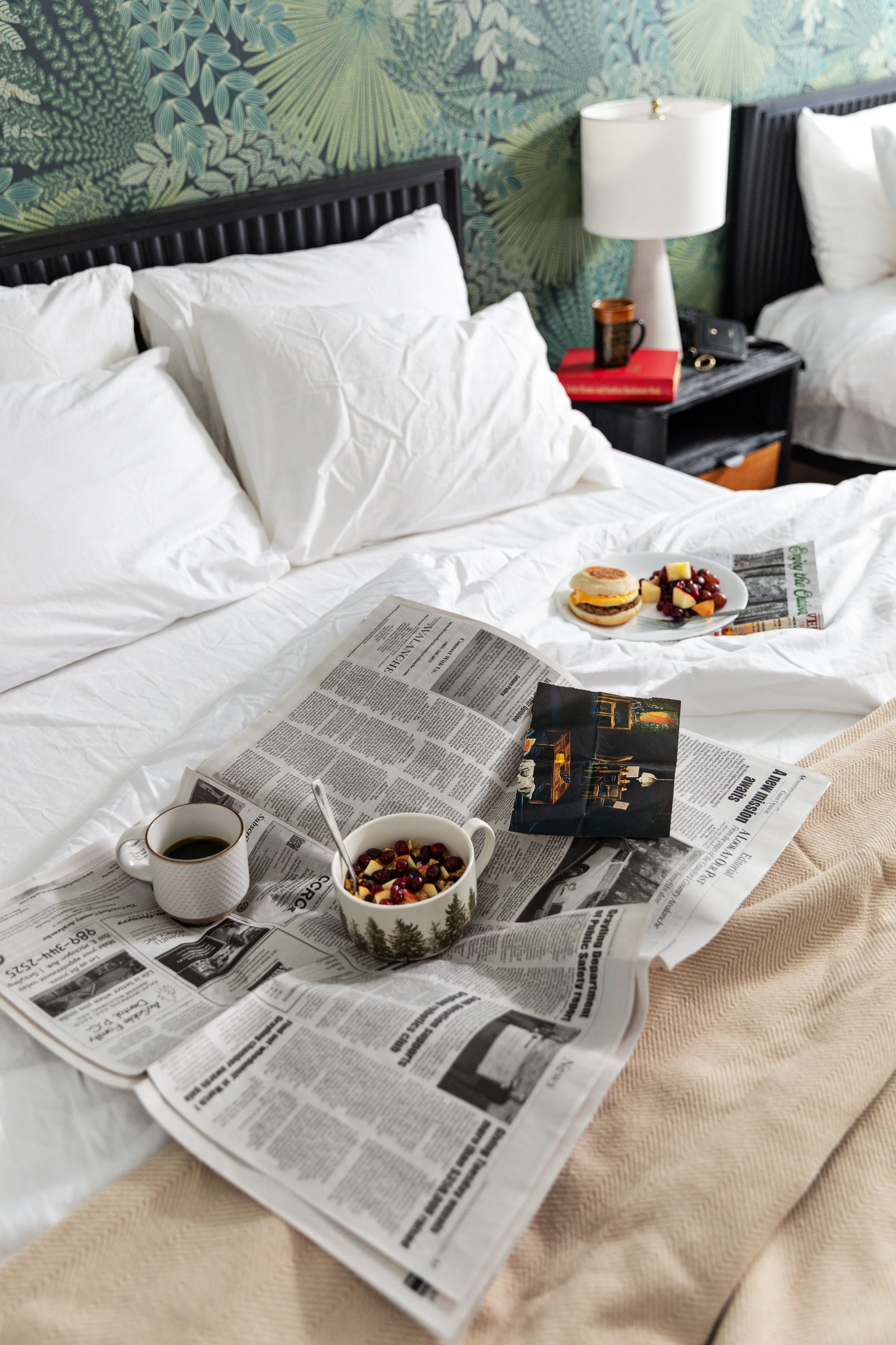 A hotel bed with white bedding and pillows, with a newspaper, a cup of coffee, a bowl of breakfast cereal with berries, a magazine, a plate with a sandwich and fruit, and a spoon on the bed. There is a black nightstand with a lamp, a mug, a book, and