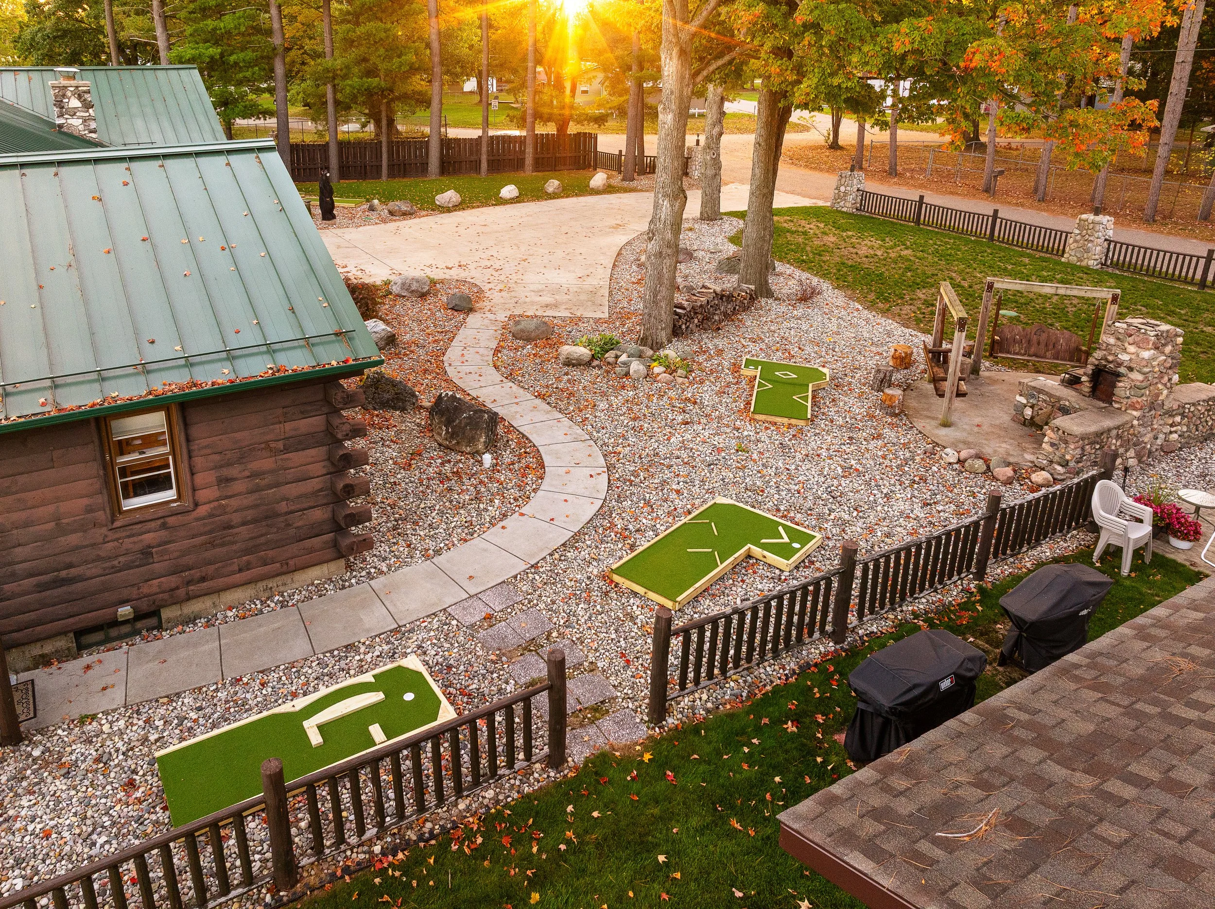 A backyard with a wooden house, a stone pathway, and a gravel yard featuring two shuffleboard courts. There are tall trees, a stone firepit with a wooden bench, outdoor chairs, and a fence. The sun is setting in the background, casting warm light ove