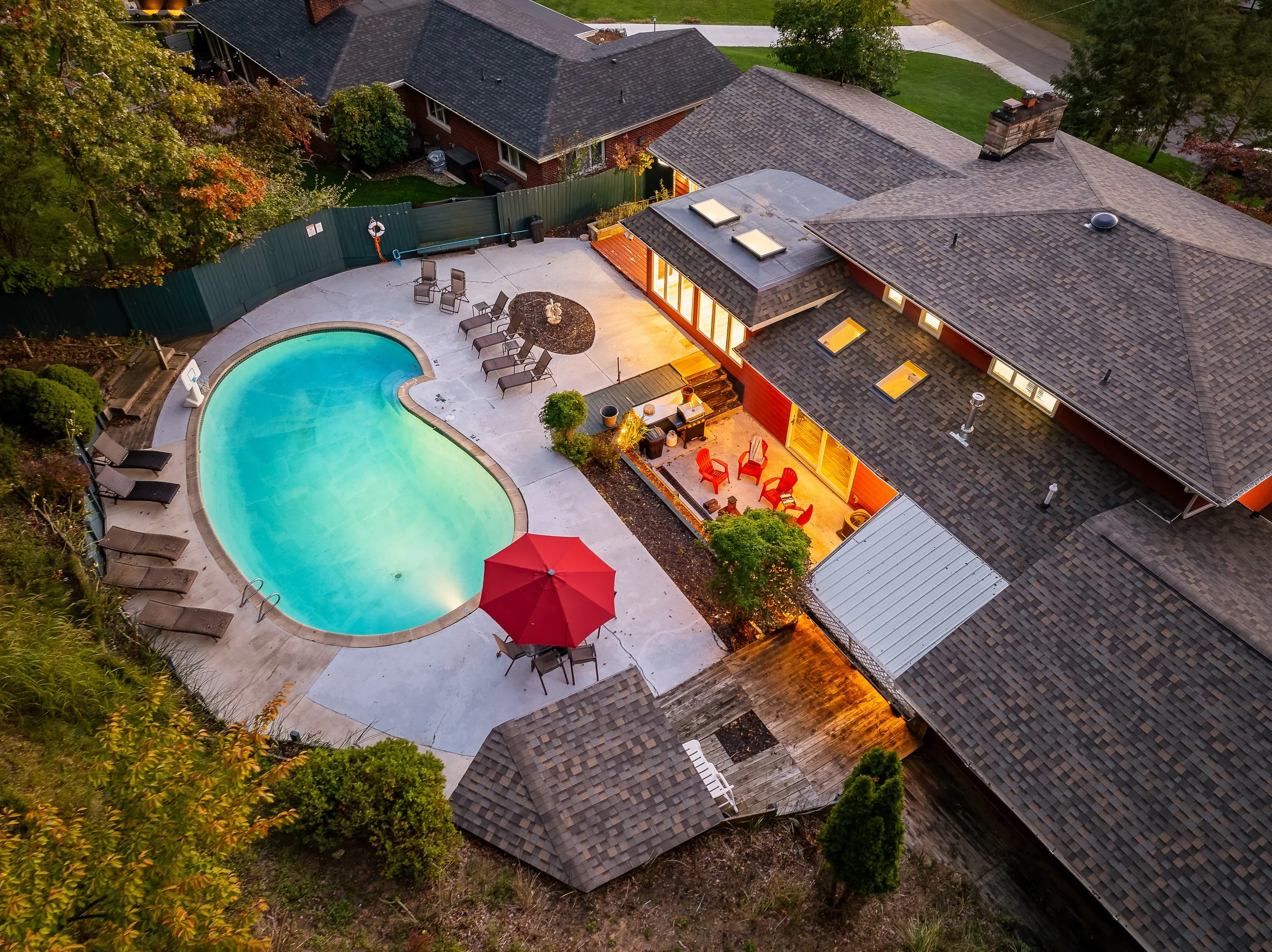 Aerial view of a backyard with a kidney-shaped swimming pool, surrounded by outdoor furniture, a house with a covered porch illuminated at night, trees, and neighboring houses.