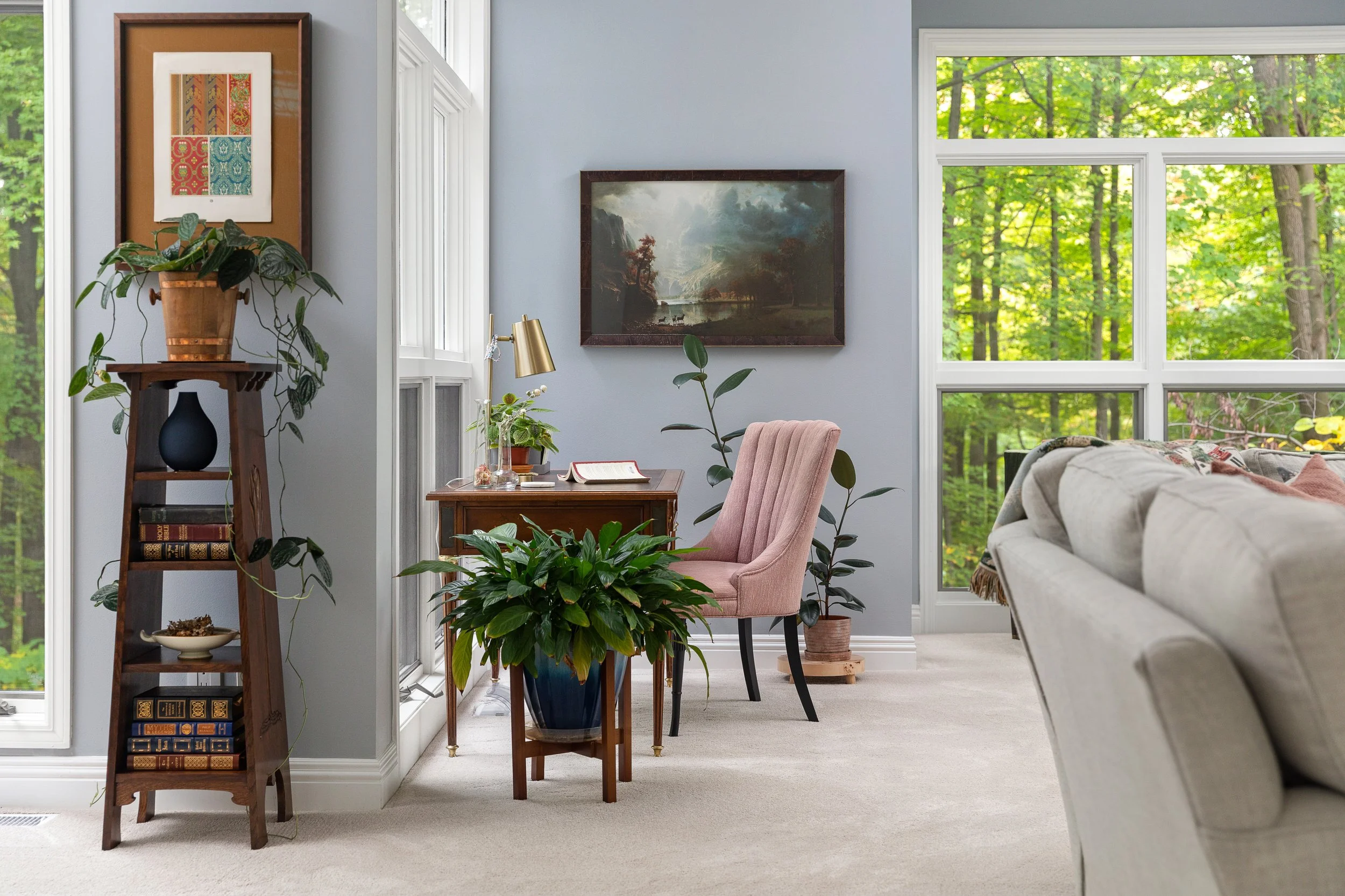 A cozy living room with large windows overlooking a green, leafy forest. There's a small wooden desk with a pink upholstered chair, a brass desk lamp, and plants. A framed painting hangs on the light gray wall. A beige couch is partially visible on t