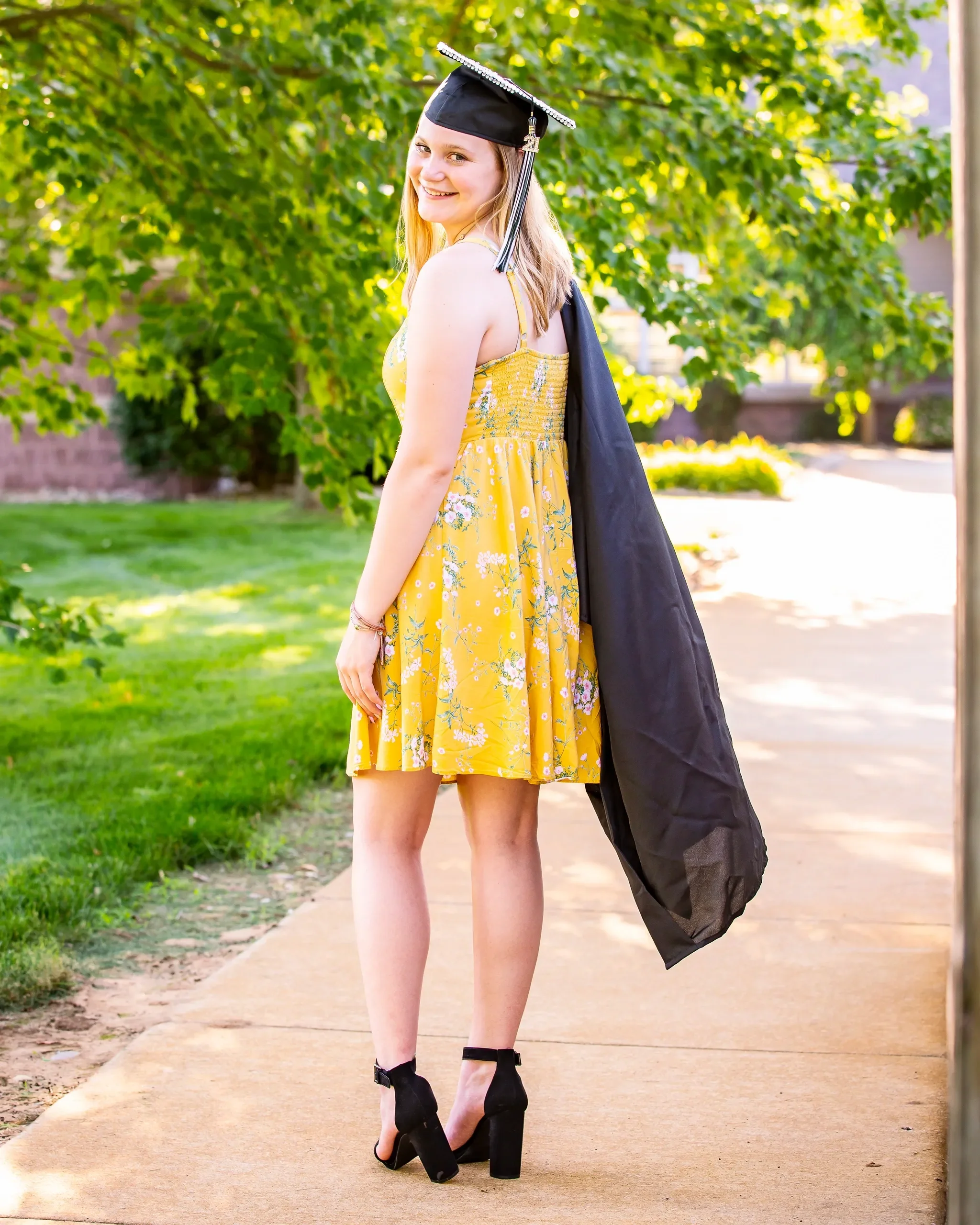 Young woman in a yellow floral dress and black high heels holding a graduation gown and cap, standing on a sidewalk in a park with trees and grass, smiling at the camera.