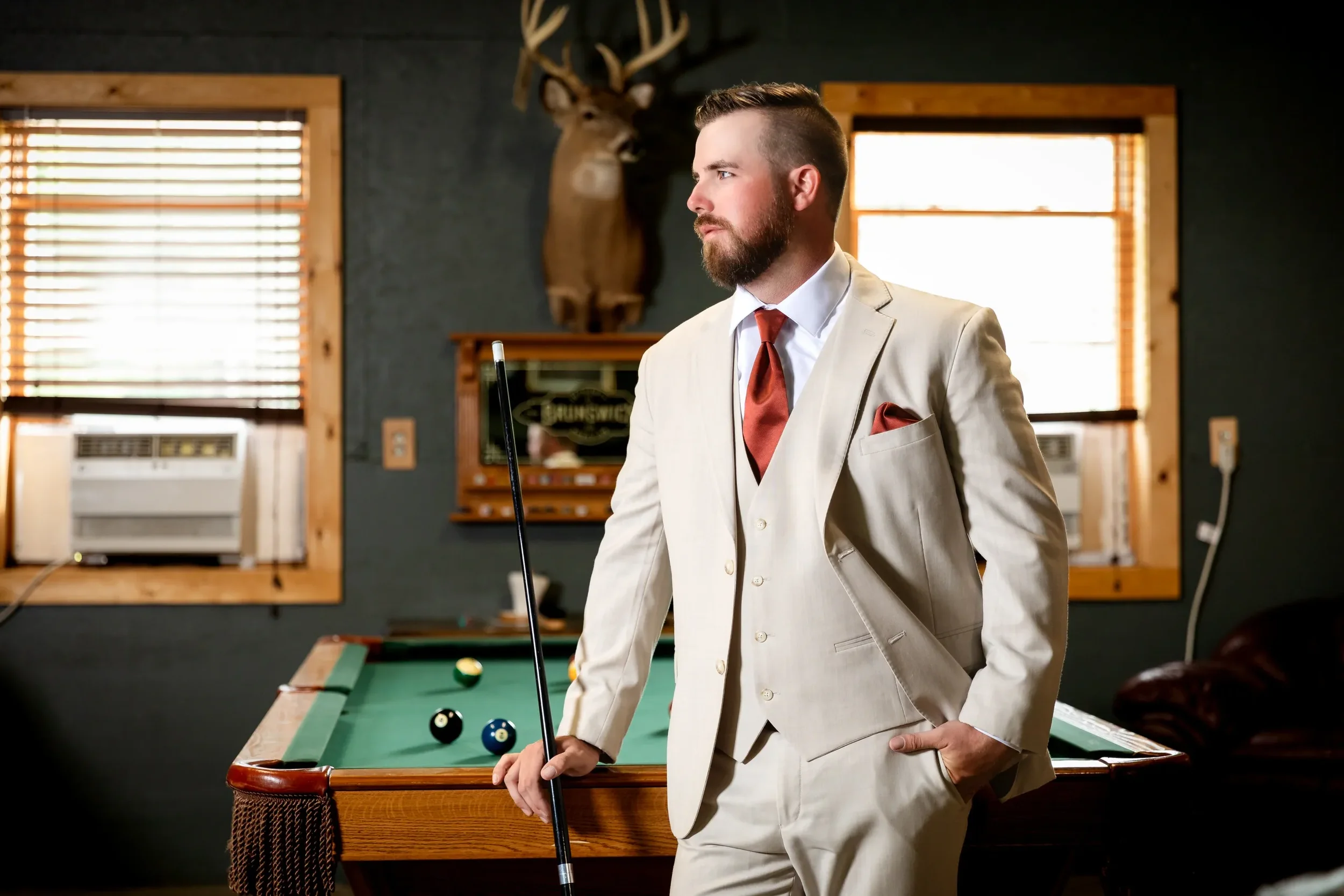A man in a cream-colored suit holding a pool cue standing next to a pool table in a room with wood window frames and a mounted deer head.