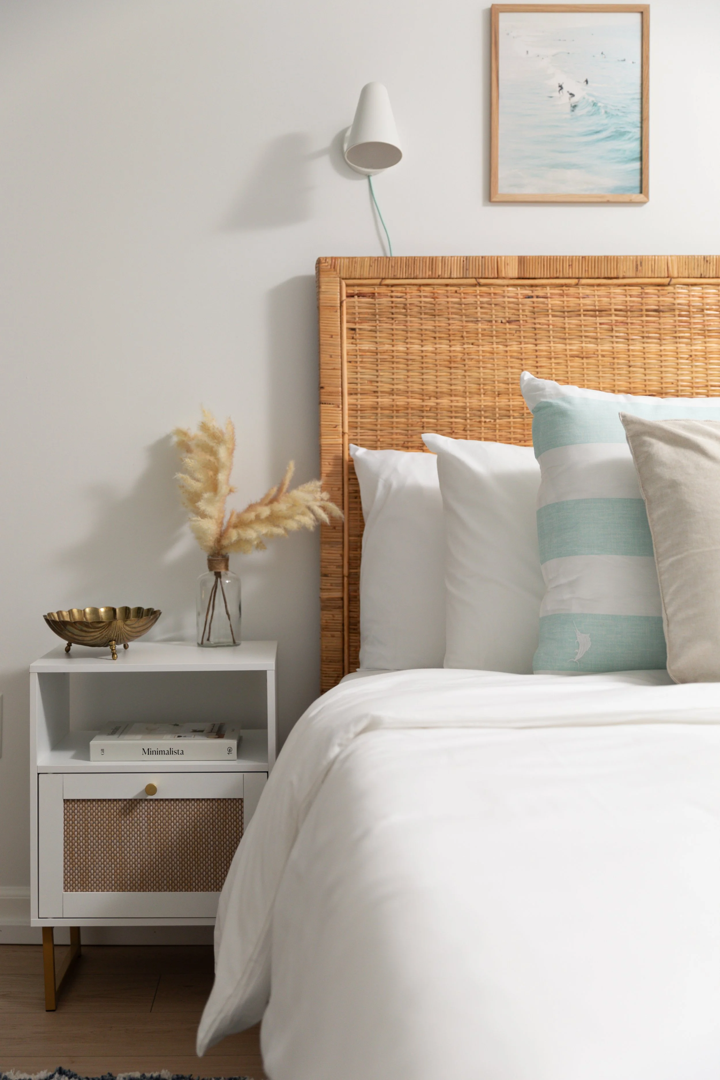 A cozy bedroom with a rattan headboard, white bedding, and decorative pillows, a white nightstand with a glass vase of dried pampas grass and a gold dish, and wall art depicting waves and surfers, all in a light, neutral color palette.