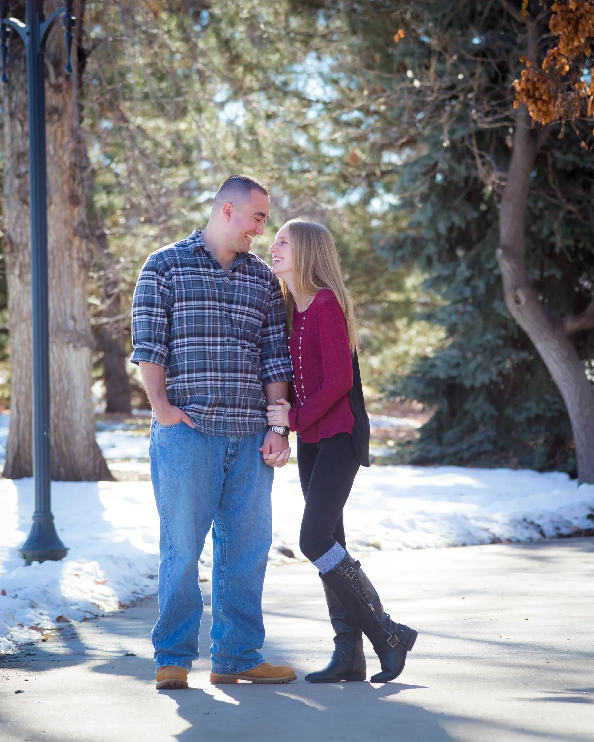 A happy couple holding hands and smiling while walking outdoors in a park during winter, with snow on the ground and autumn trees in the background.