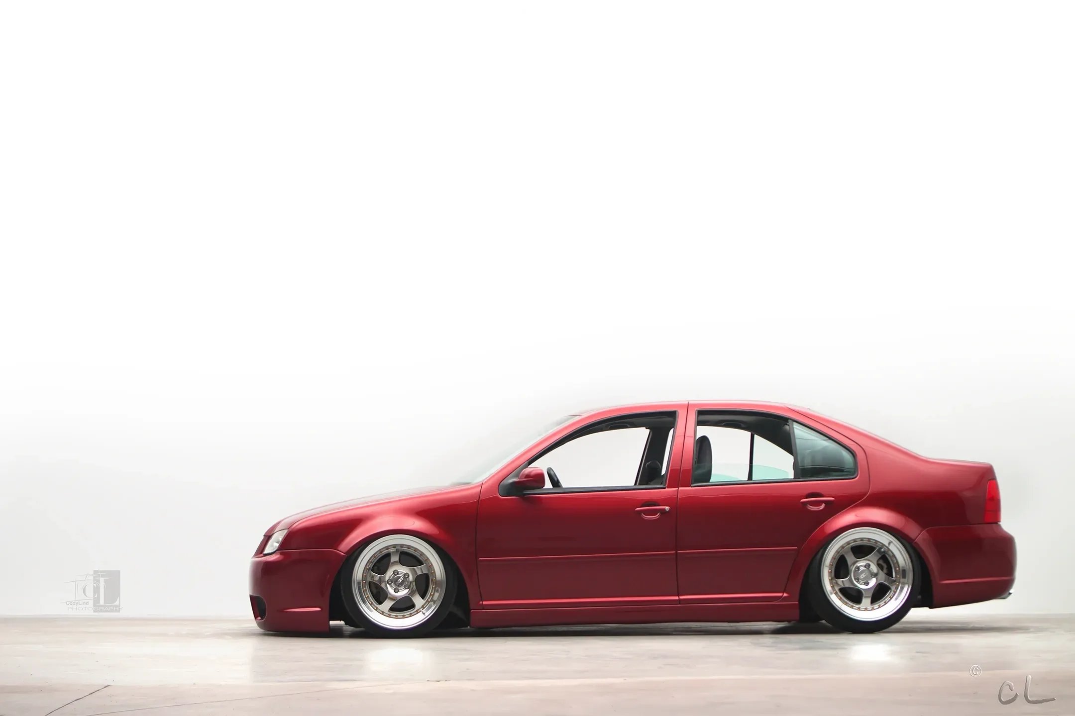 Red lowered sedan with custom wheels parked on a smooth concrete floor against a plain white background
