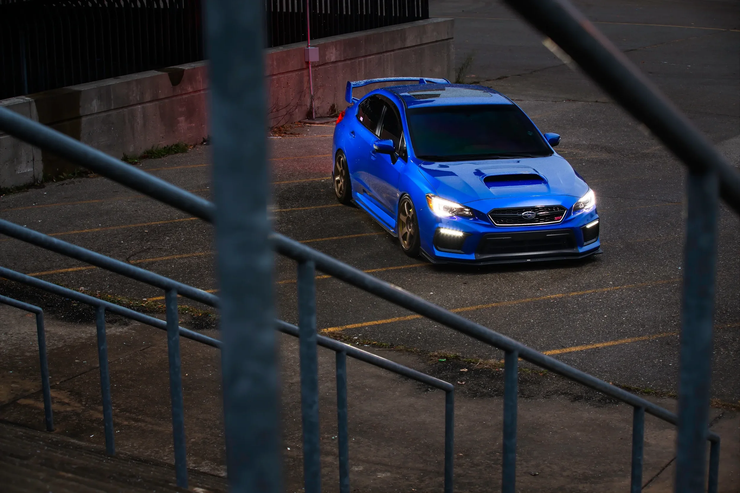Blue Subaru car parked in an empty parking lot at night, viewed through metal railings.